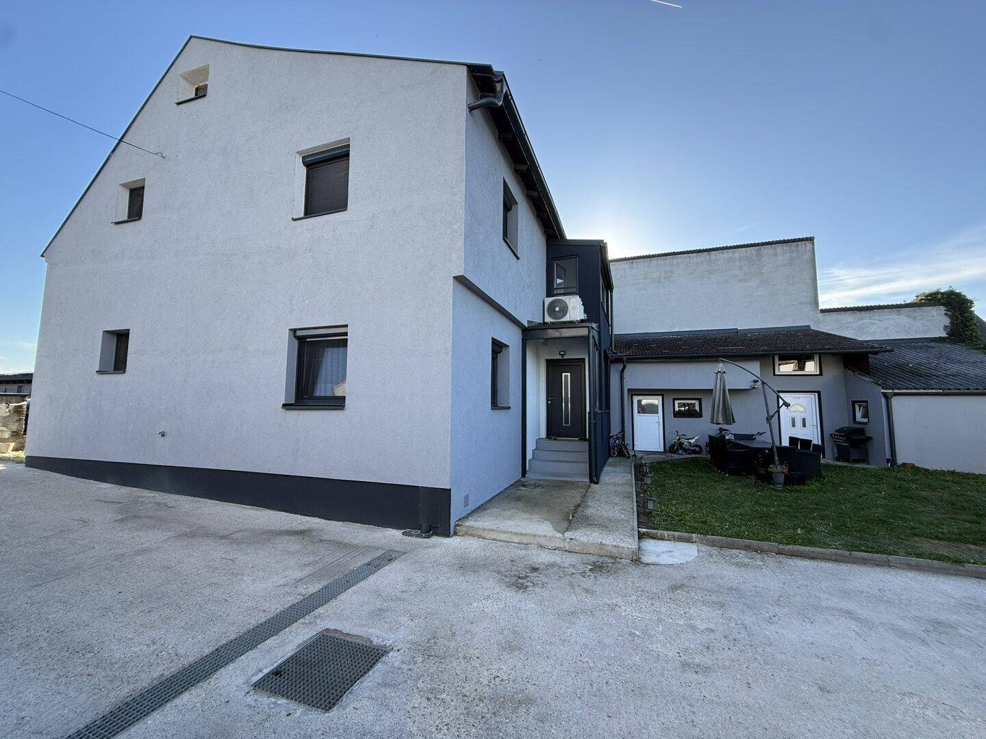 Modernes Haus mit grauer Fassade, kleinen Fenstern und einem gepflasterten Eingangsbereich.