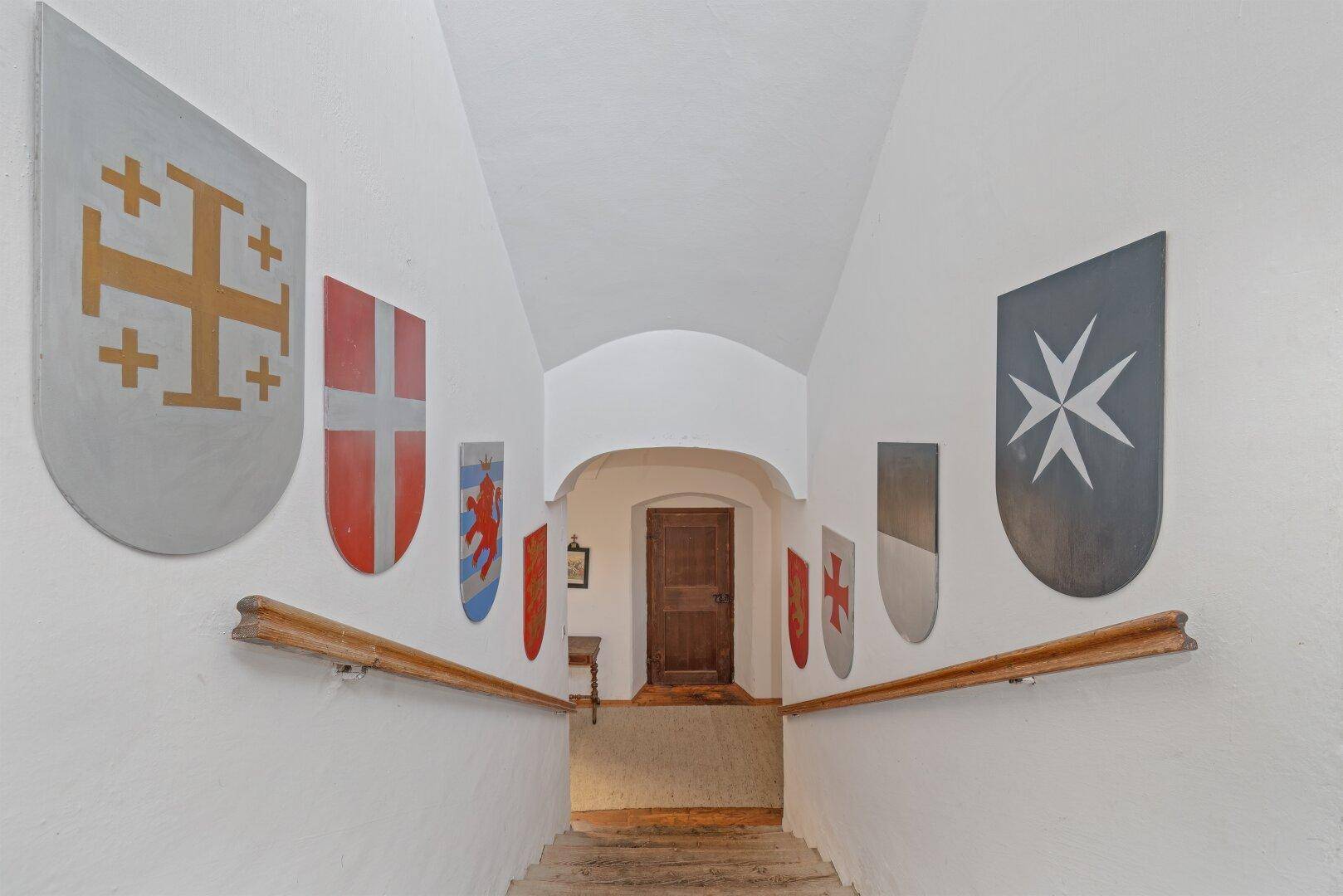 Treppenaufgang mit Wappen an den Wänden und hölzernen Handläufen in einem historischen Gebäude.