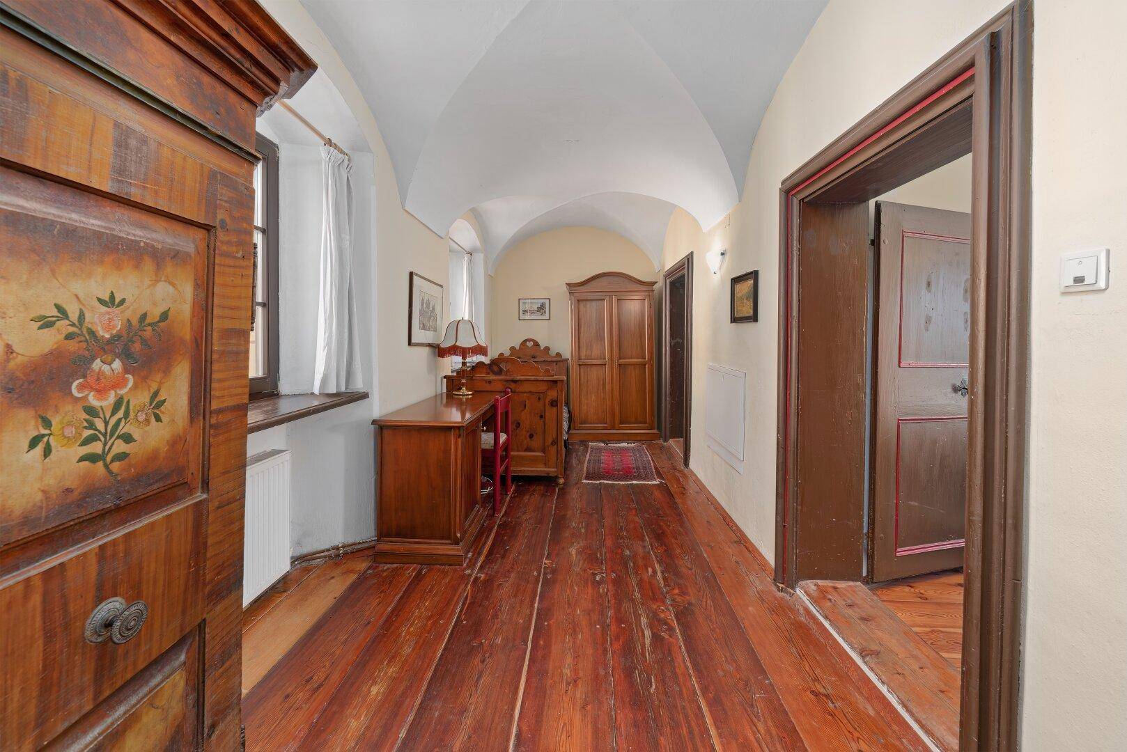 Langer Flur mit Holzdielenboden, Gewölbedecke und antiken Holzmöbeln, führt zu weiteren Räumen.