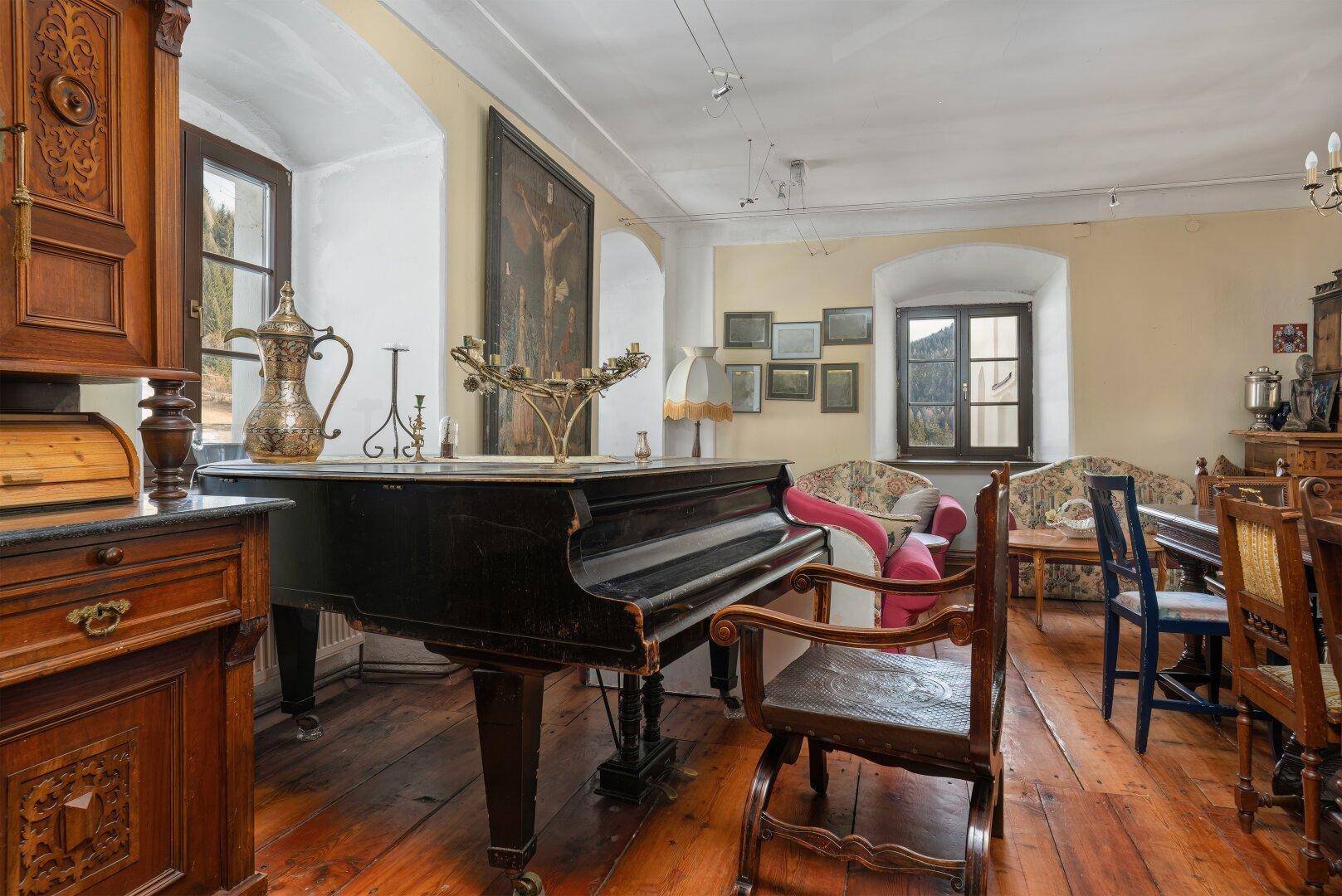 Geräumiges Wohnzimmer mit Holzdielenboden, Flügel und antiken Möbeln, durch Fenster hell beleuchtet.