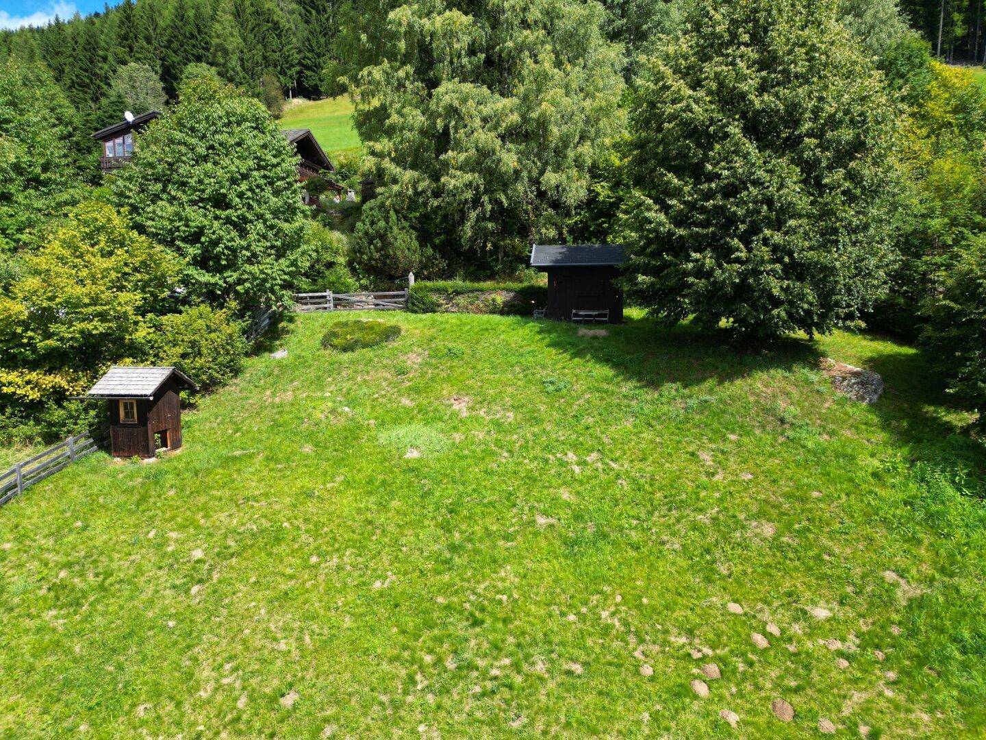 Grüne Wiese mit kleinen Holzhütten und Bäumen in einer ländlichen Umgebung unter hellem Himmel.