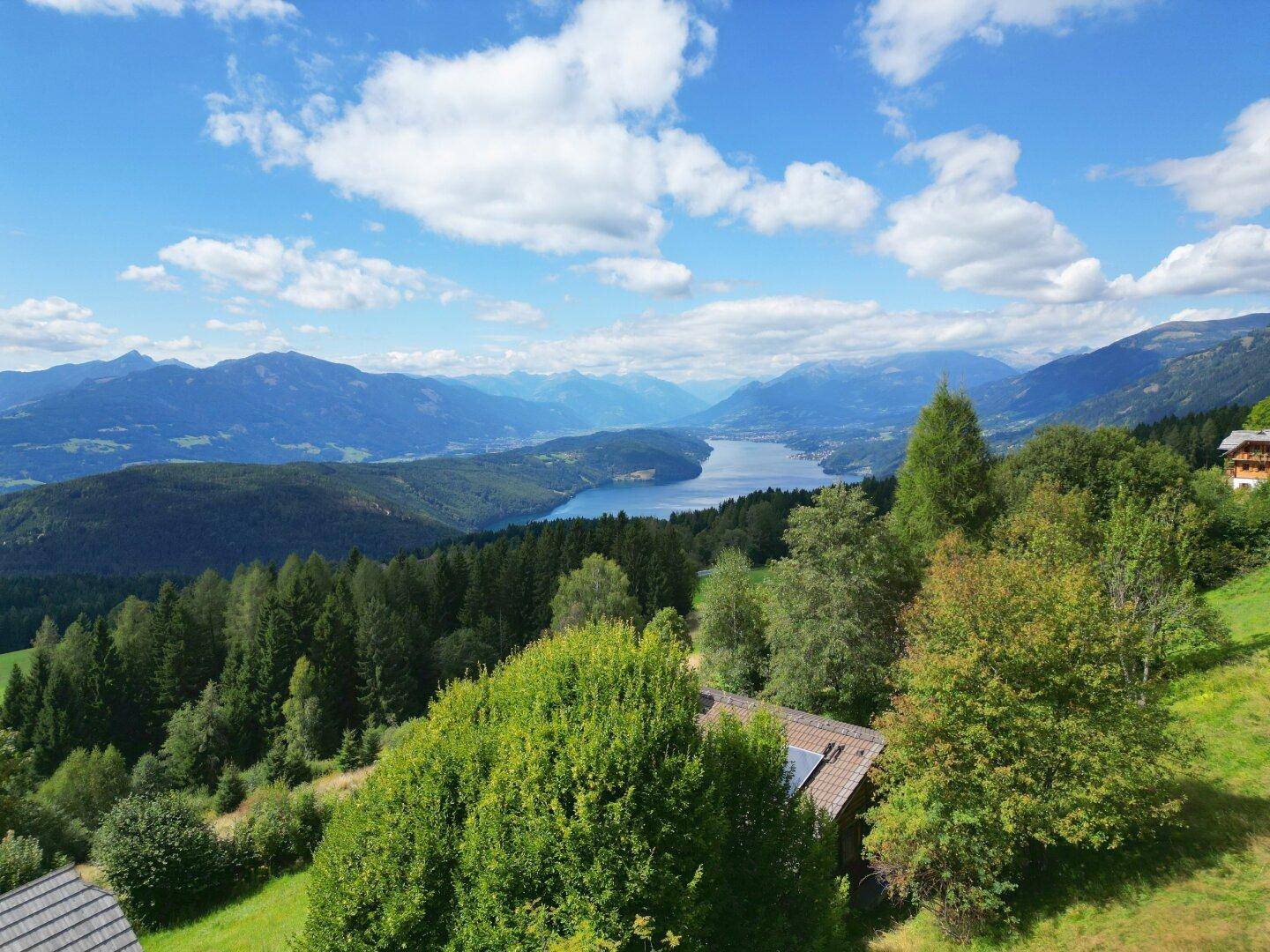 Weitläufiger Blick auf ein Haus mit Solaranlage, See und Bergpanorama unter blauem Himmel.