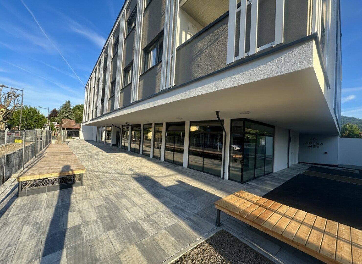 Seitenansicht eines modernen Gebäudes mit großen Glasfronten im Erdgeschoss und gepflastertem Außenbereich bei Sonnenschein.