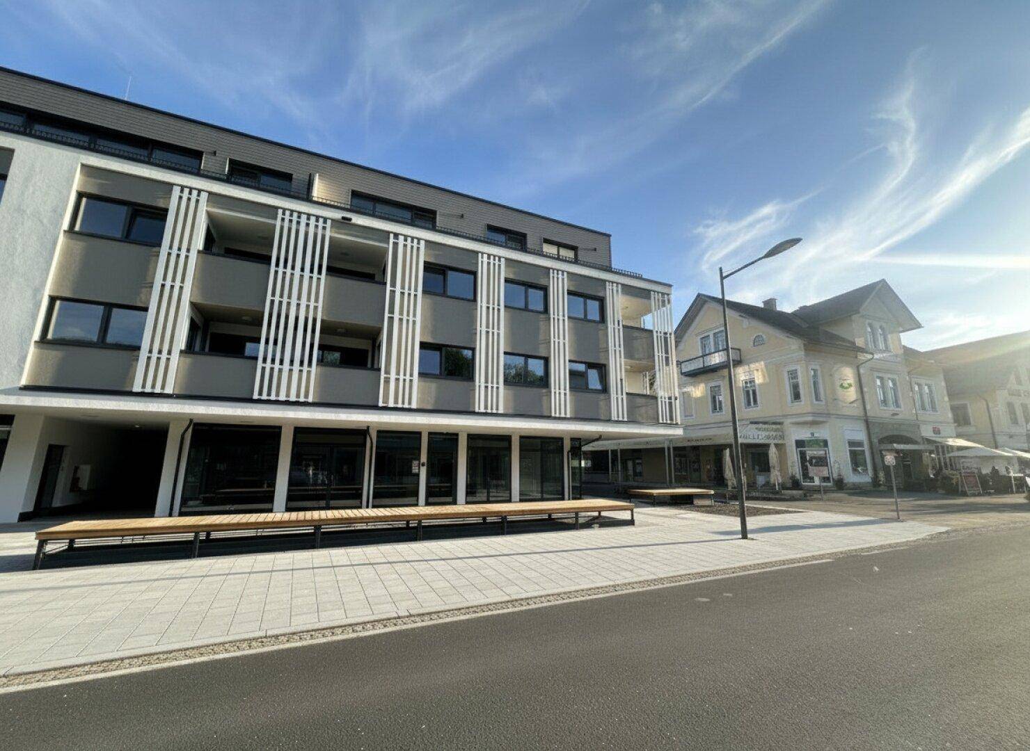 Ansicht eines modernen Wohn- und Geschäftshauses mit Balkonen, großen Fenstern und Sitzgelegenheiten im Außenbereich bei hellem Wetter.