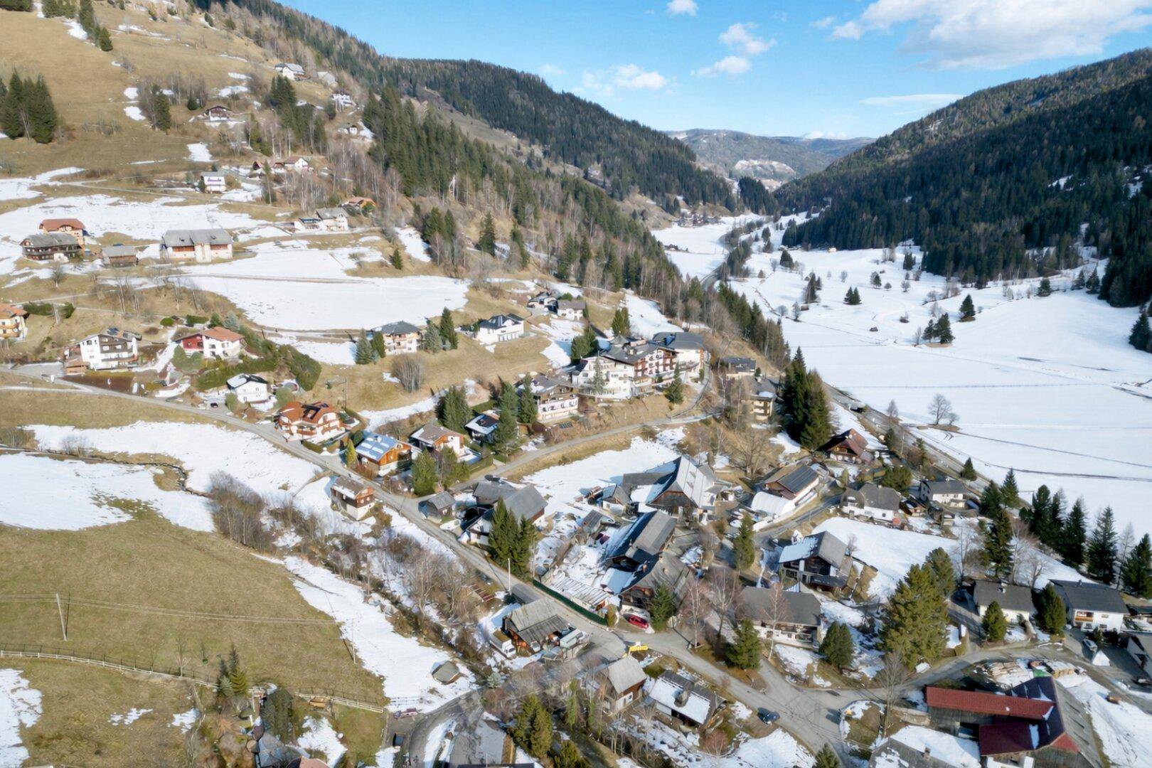 Panoramische Luftaufnahme eines malerischen Tals mit einem Dorf, umgeben von schneebedeckten Hängen und Wäldern.