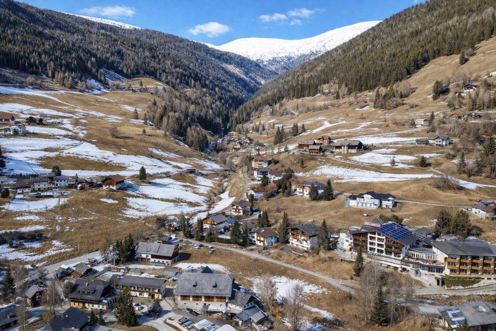 Weitläufige Luftaufnahme eines alpinen Tals mit einem Dorf, umgeben von schneebedeckten Bergen und Wäldern.