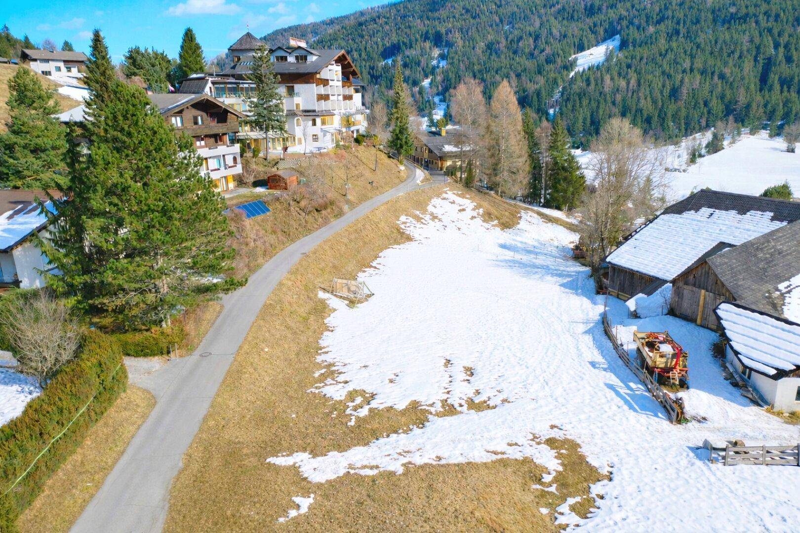 Blick aus der Vogelperspektive auf eine Straße, Häuser am Hang und ein schneebedecktes Feld mit Bergen im Hintergrund.