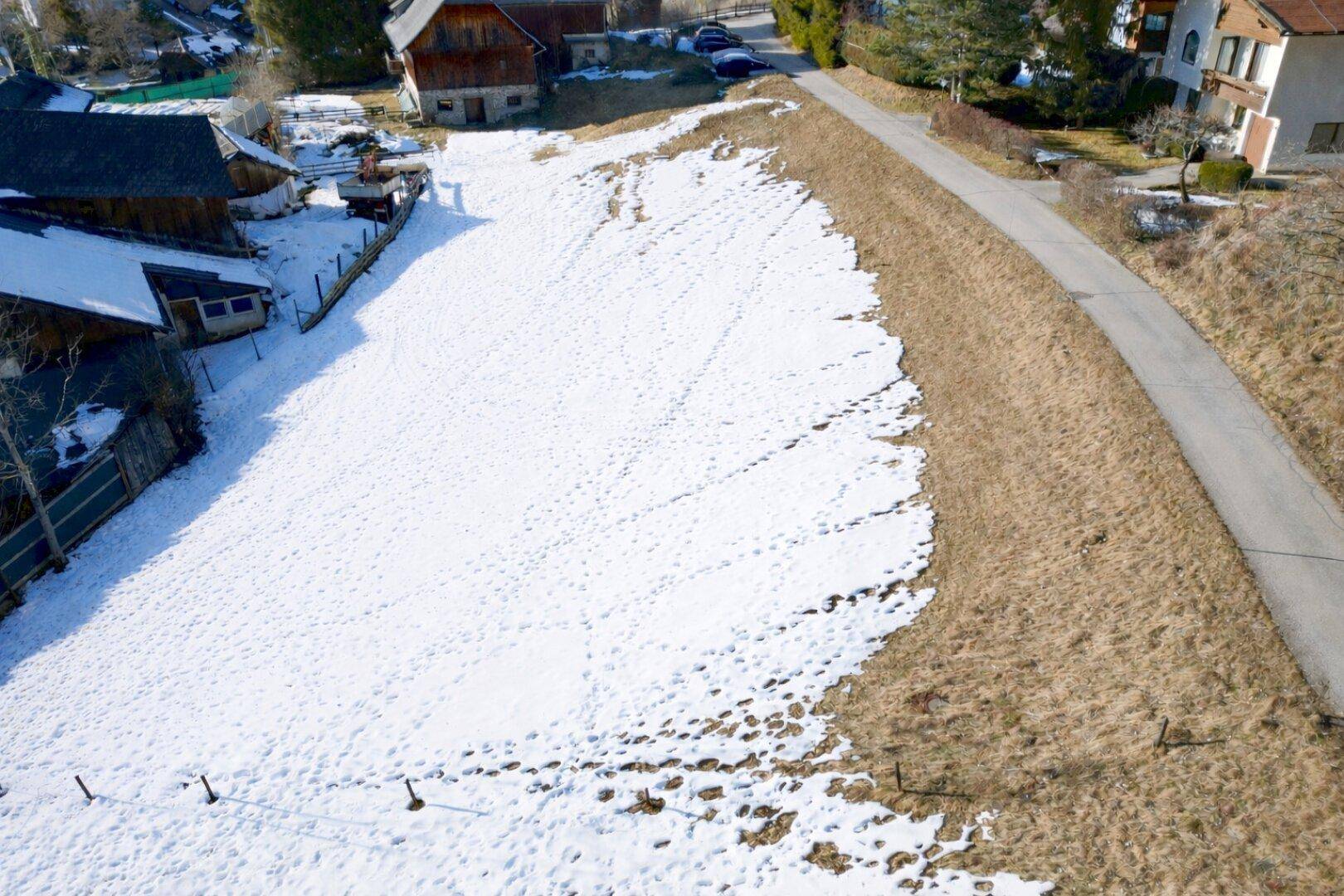 Luftaufnahme eines schneebedeckten Feldes mit Fußspuren, einer Straße und ländlichen Gebäuden im Hintergrund.