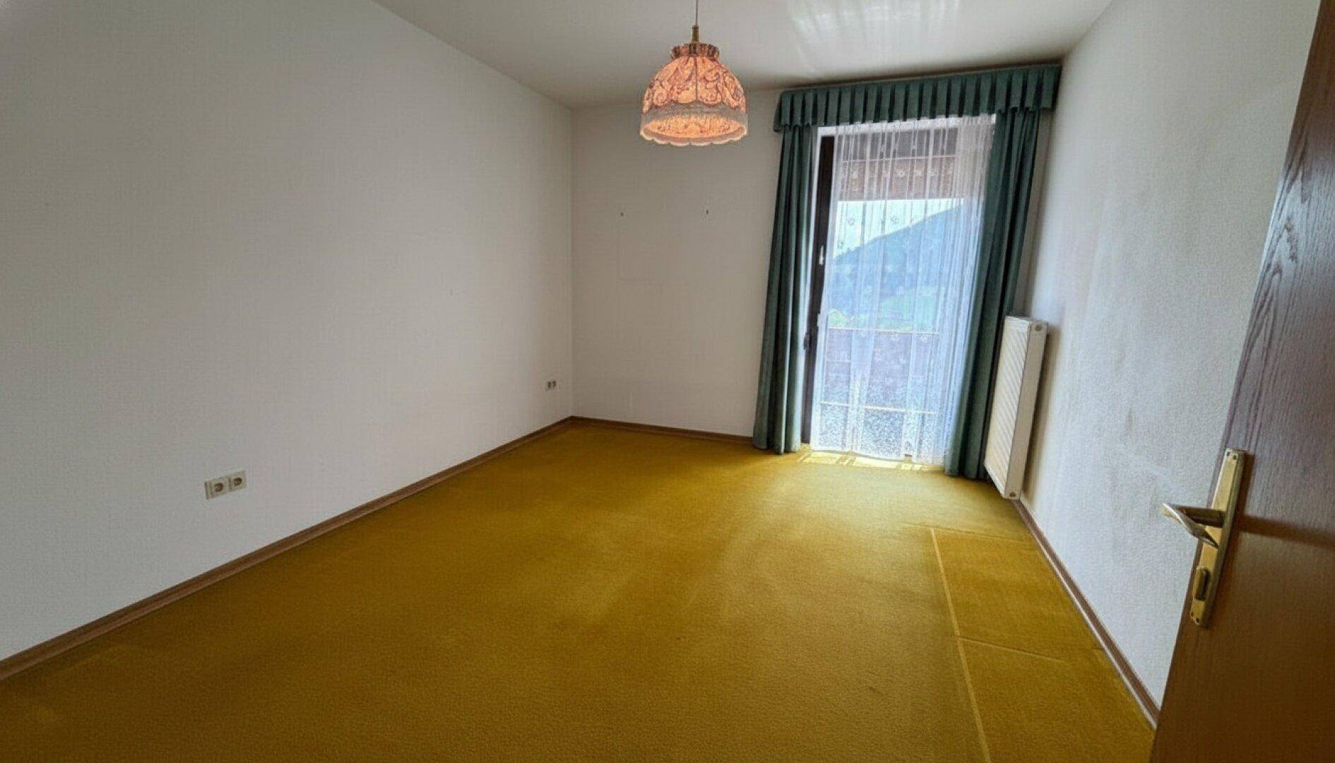 Leeres Zimmer mit gelbem Teppichboden, weißen Wänden und einem Fenster mit Zugang nach draußen.