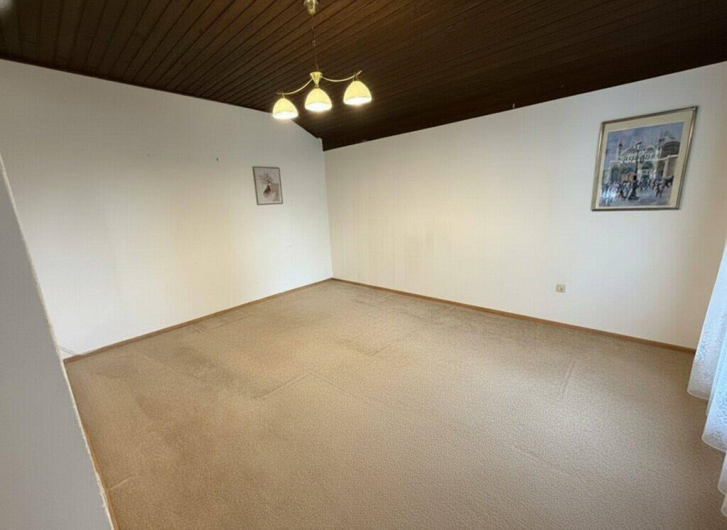 Geräumiges Zimmer mit beigem Teppichboden, weißen Wänden und einer dunklen Holzdecke.