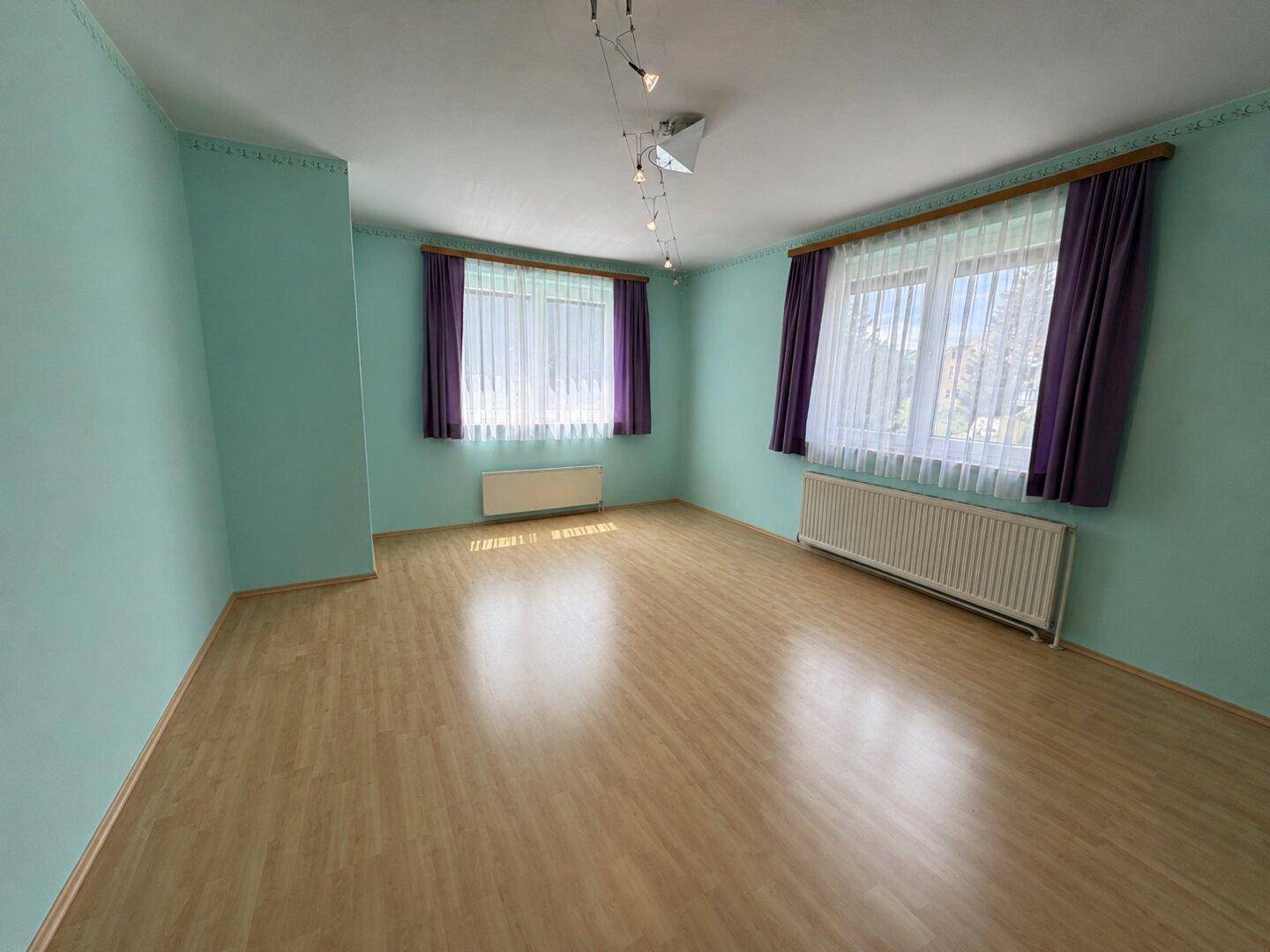 Helles Zimmer mit Laminatboden, türkisfarbenen Wänden und zwei Fenstern, die den Raum erhellen.