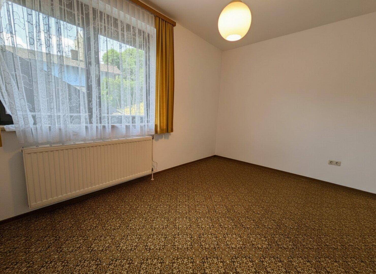 Leeres Zimmer mit gemustertem Teppichboden, weißen Wänden und einem großen Fenster für Tageslicht.
