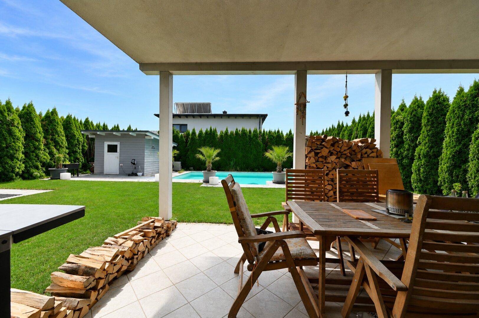 Überdachte Terrasse mit Holztisch, Sitzgelegenheiten und Blick auf den Garten und Pool.