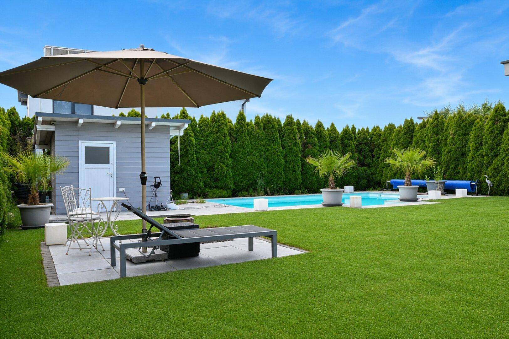 Gepflegter Garten mit Rasenfläche, Pool im Hintergrund und gemütlicher Liegefläche.