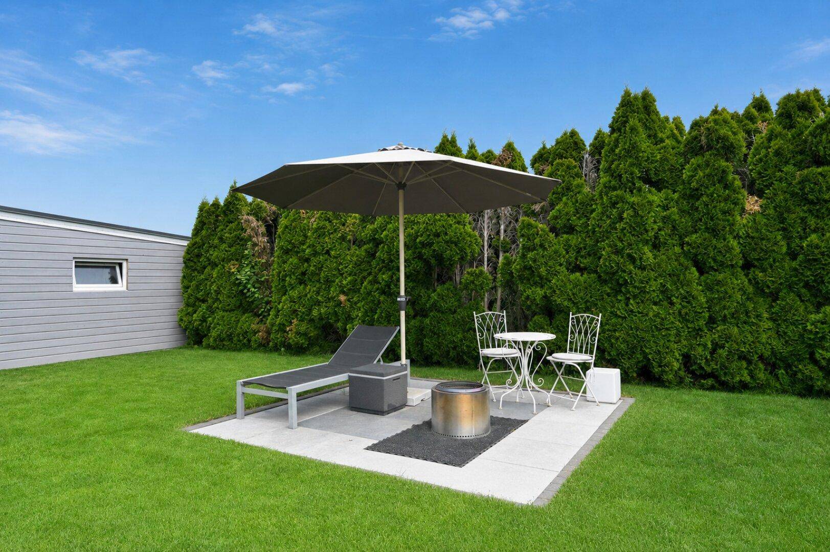 Gemütliche Terrasse mit Liegestuhl, Sonnenschirm und Feuerstelle im Garten.