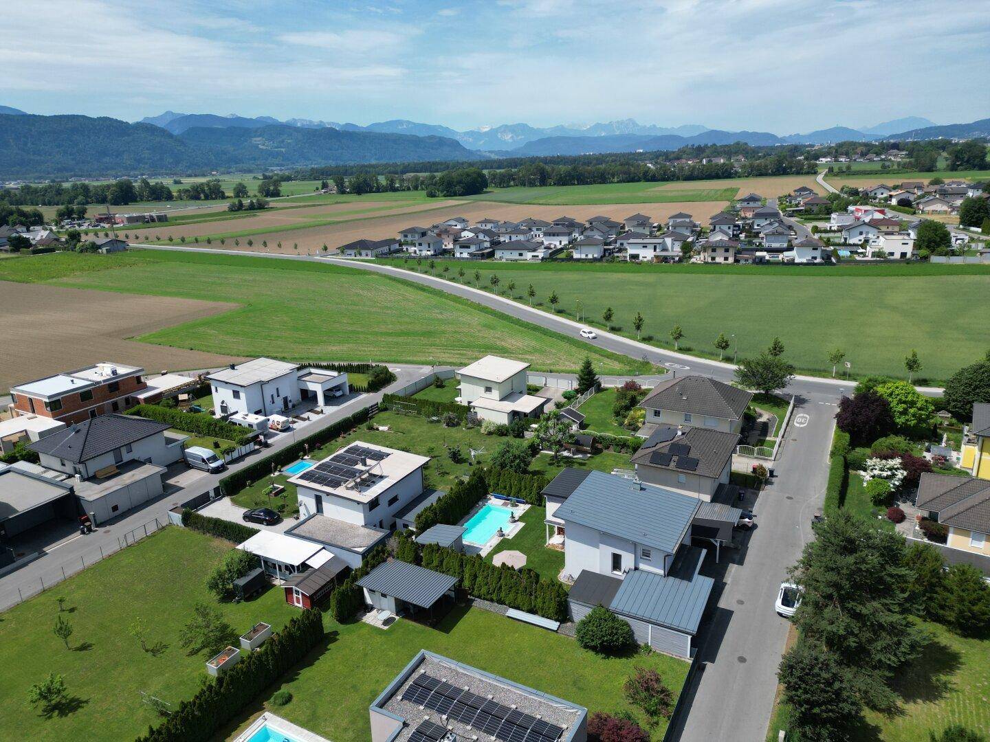 Luftaufnahme einer Wohnsiedlung mit Häusern, grünen Feldern und Bergen im Hintergrund.