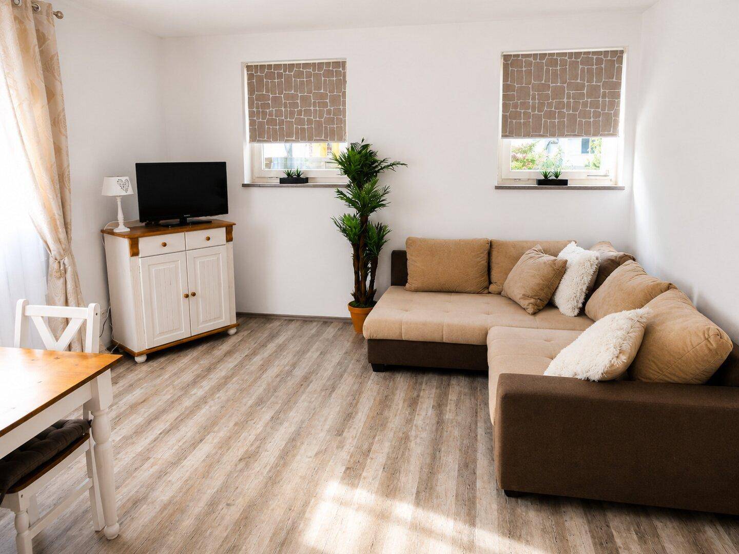 Gemütlicher Wohnbereich mit beigem Ecksofa, Fernseher auf einem weißen Schrank und viel Tageslicht.