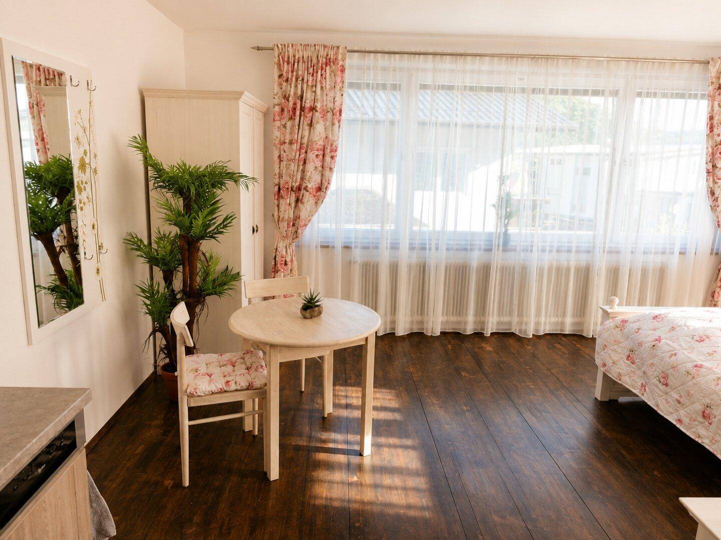 Helles Schlafzimmer mit Holzboden, großem Fenster und einem kleinen runden Esstisch.