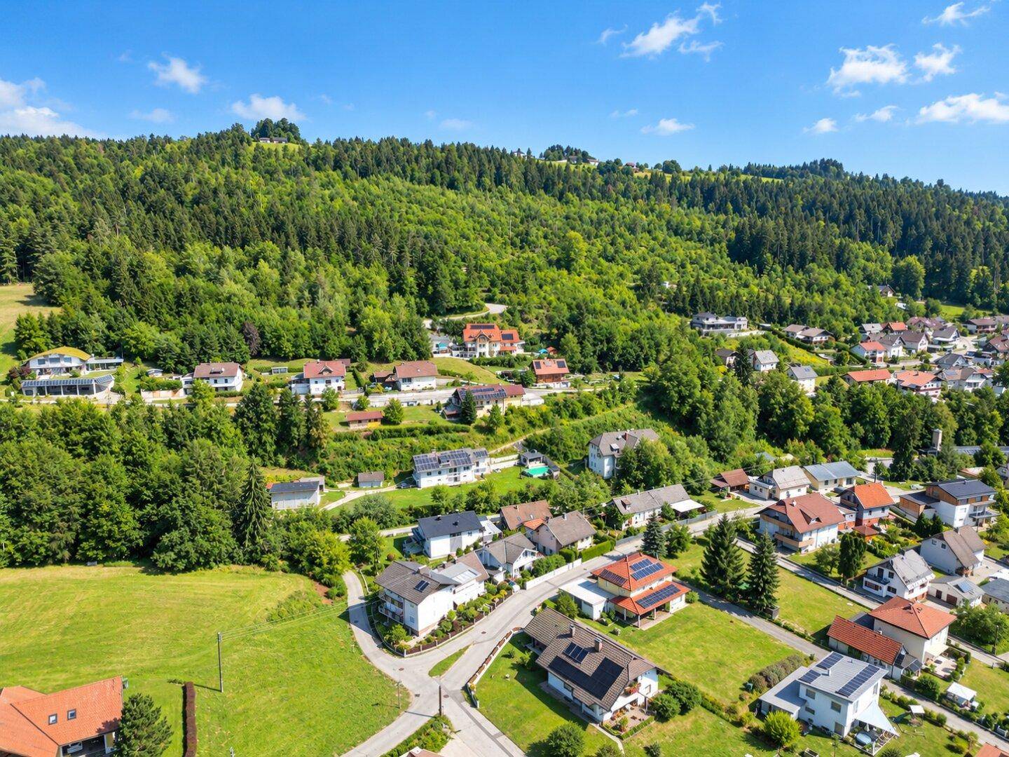 Luftaufnahme einer Wohnsiedlung eingebettet in eine grüne, bewaldete Hügellandschaft unter blauem Himmel.