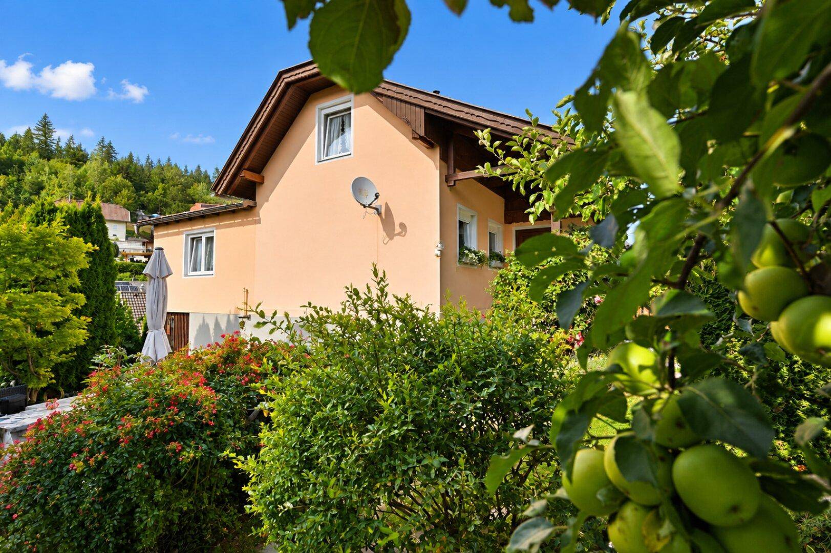 Haus umgeben von einem üppigen Garten mit Obstbäumen und grüner Vegetation unter blauem Himmel.