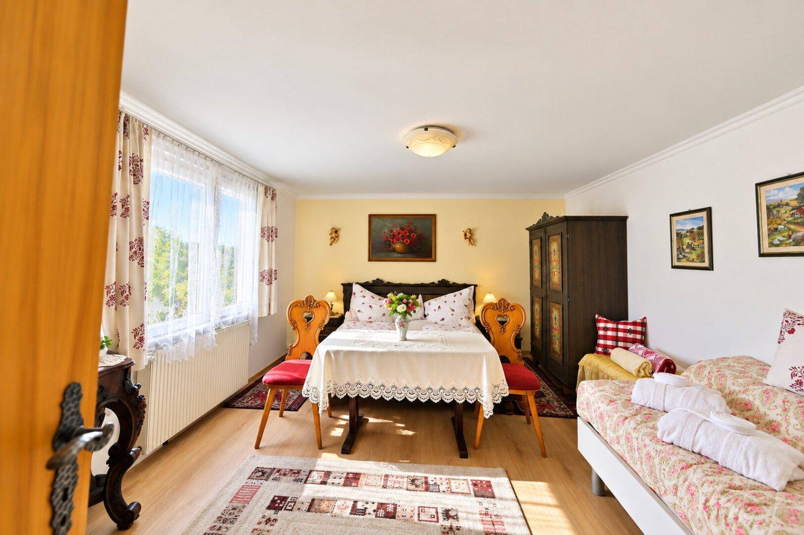 Helles Schlafzimmer mit traditionellen Möbeln, Doppelbett und einem Tagesbett, durch ein Fenster gut beleuchtet.