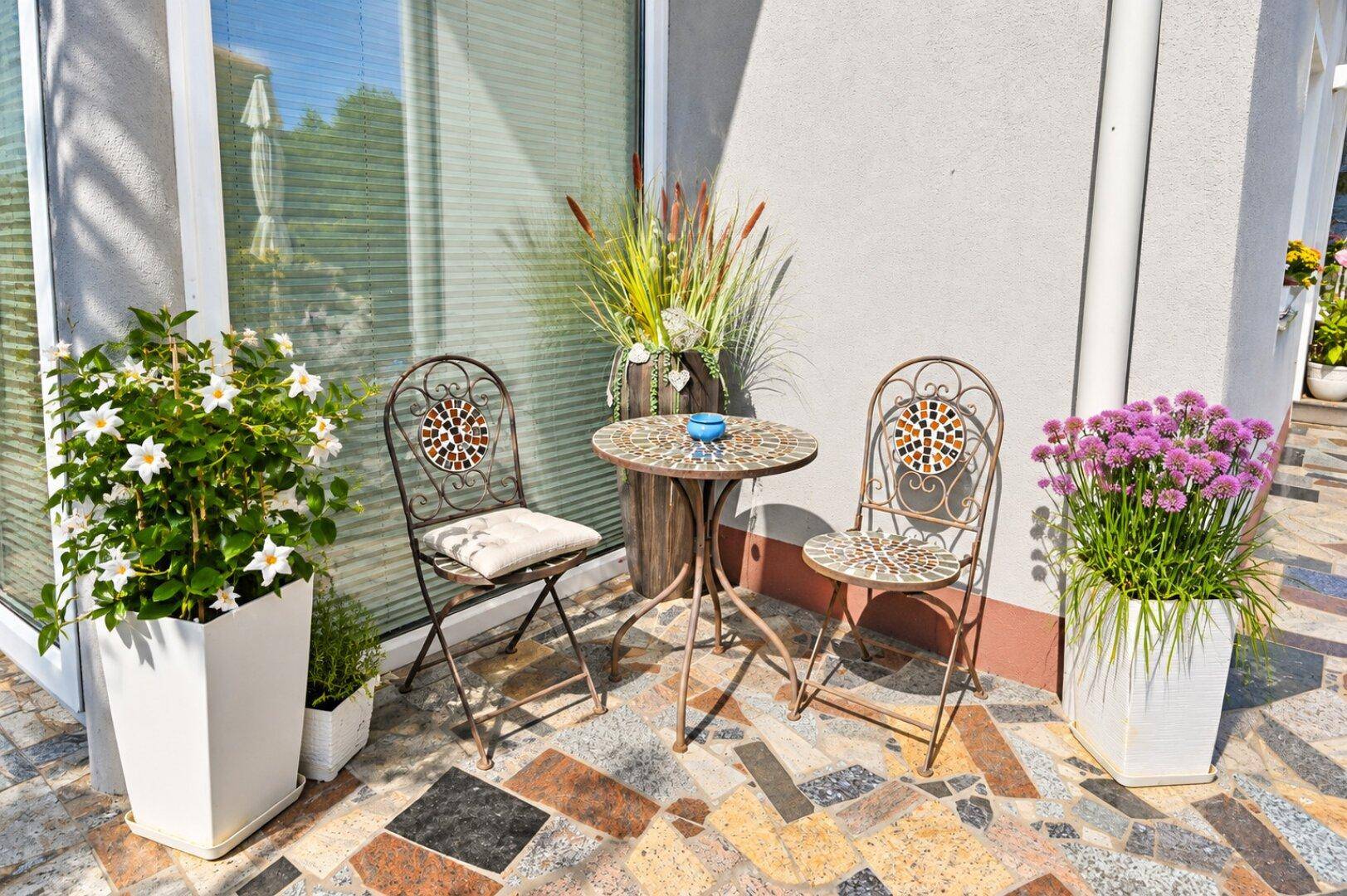 Kleine, sonnige Terrasse mit Mosaikfliesen und einem Bistrotisch mit Stühlen, umgeben von Pflanzen.