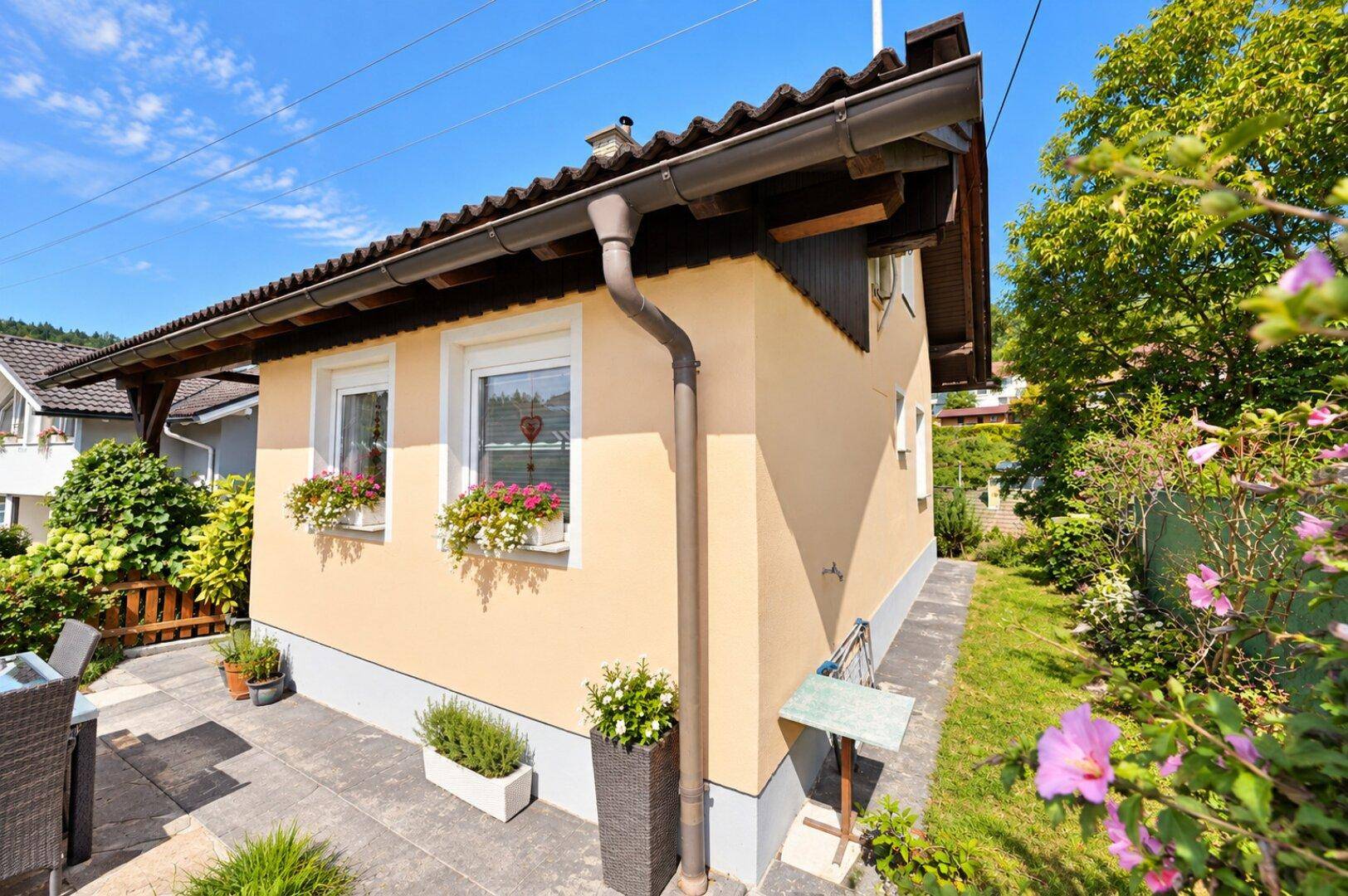 Außenansicht eines Hauses mit gepflasterter Terrasse, Gartenmöbeln und Blumenkästen an den Fenstern.