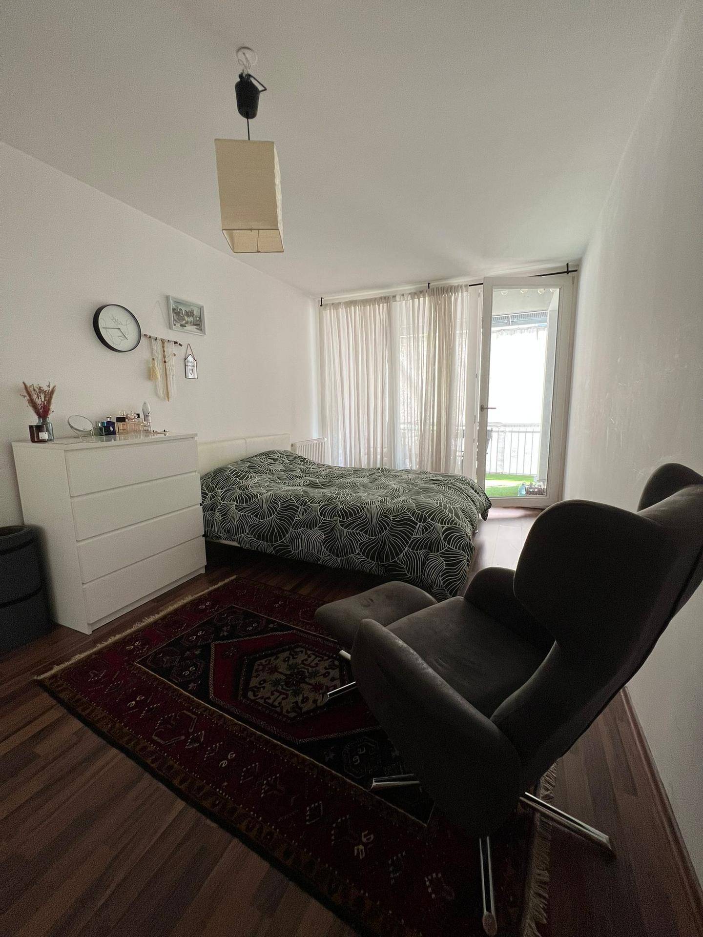 Schlafzimmer mit Holzboden, Bett, Sessel und Zugang zu einem sonnigen Balkon.