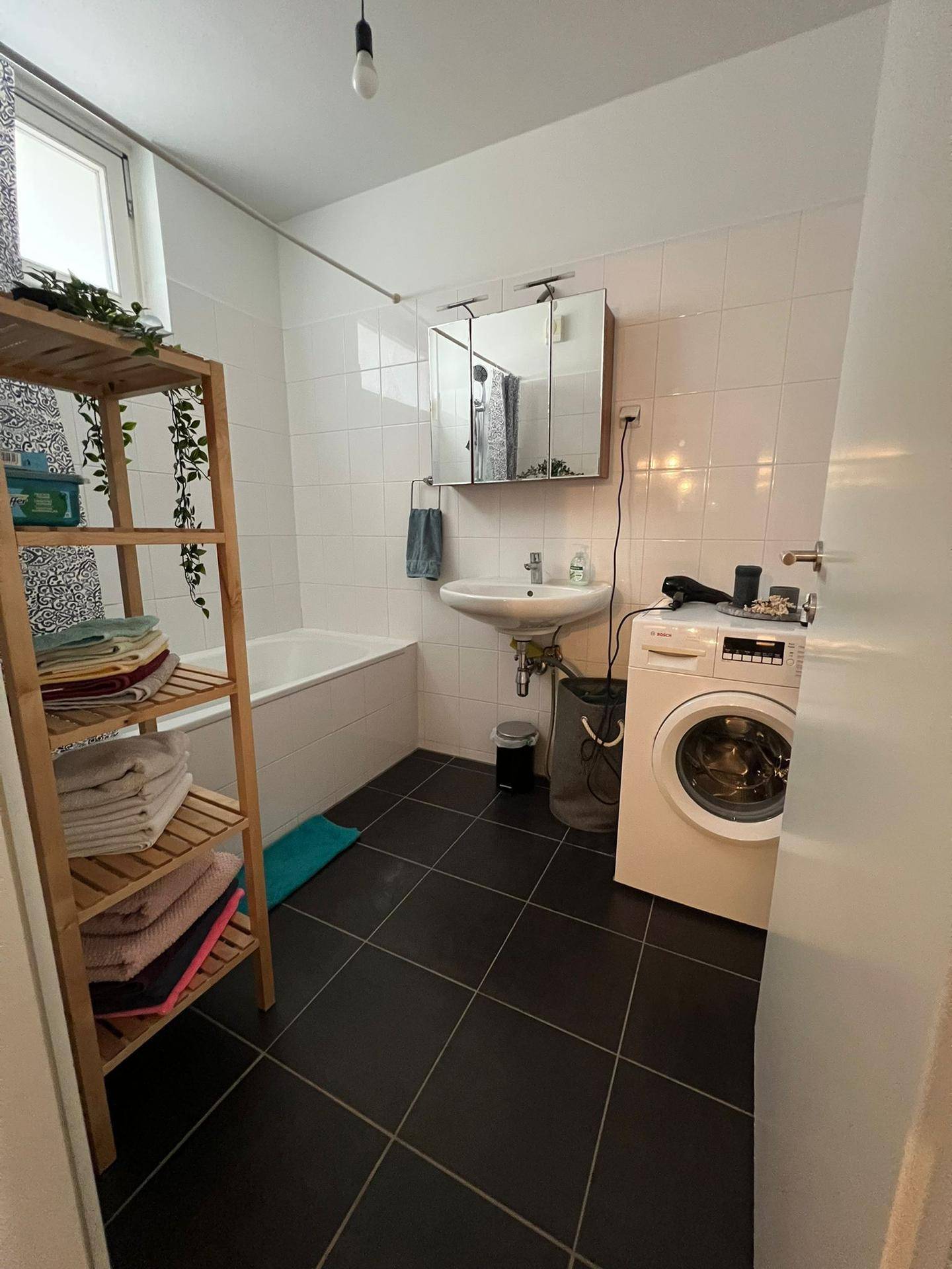 Badezimmer mit Badewanne, Waschbecken, Spiegelschrank und Waschmaschine, hell gefliest.