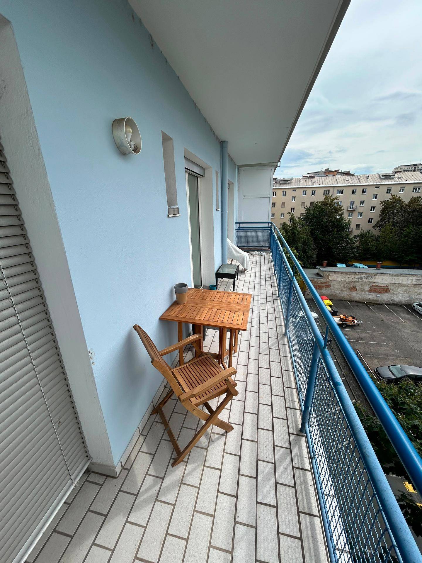 Langer Balkon mit Fliesenboden, blauer Brüstung und kleiner Sitzgelegenheit im Freien.