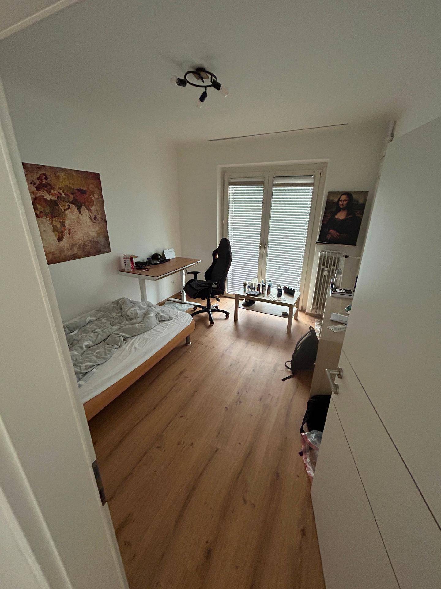 Helles Schlafzimmer mit Einzelbett, Schreibtisch und Fenster mit Jalousien.