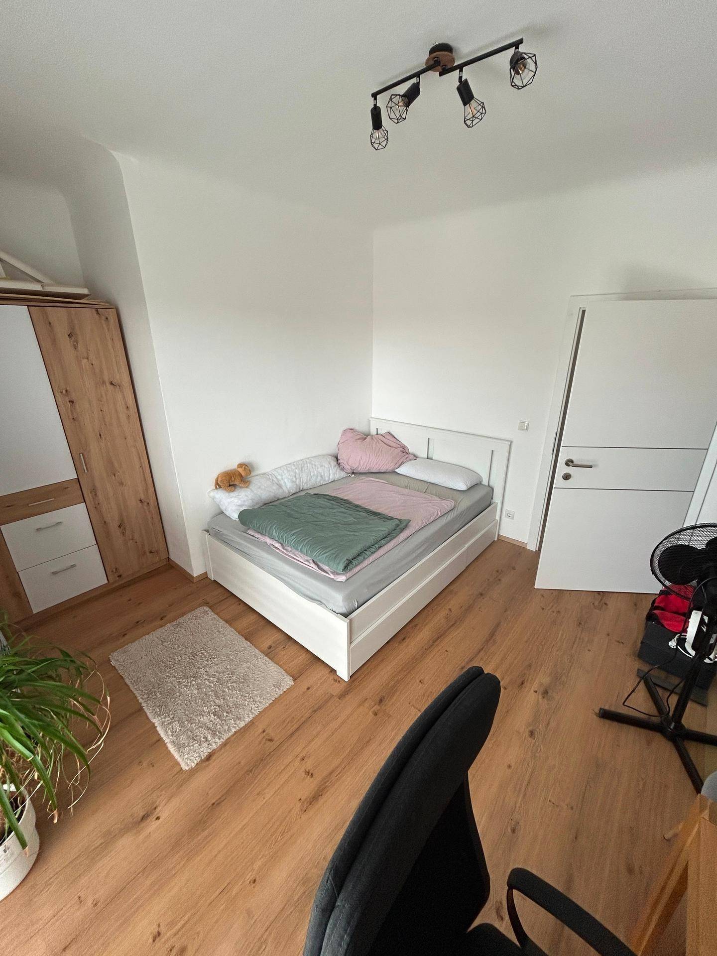 Helles Schlafzimmer mit Bett, Kleiderschrank und Ventilator, mit Holzboden.
