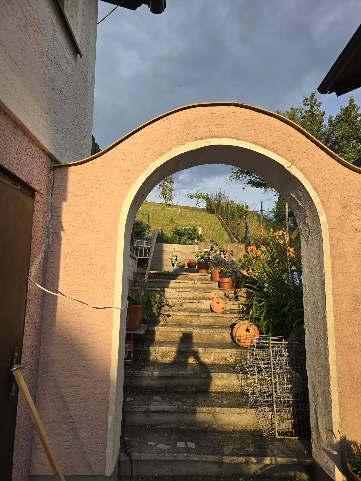 Detailansicht eines Torbogens und einer Steintreppe, die einen begrünten Hang hinaufführt, geschmückt mit Topfpflanzen.