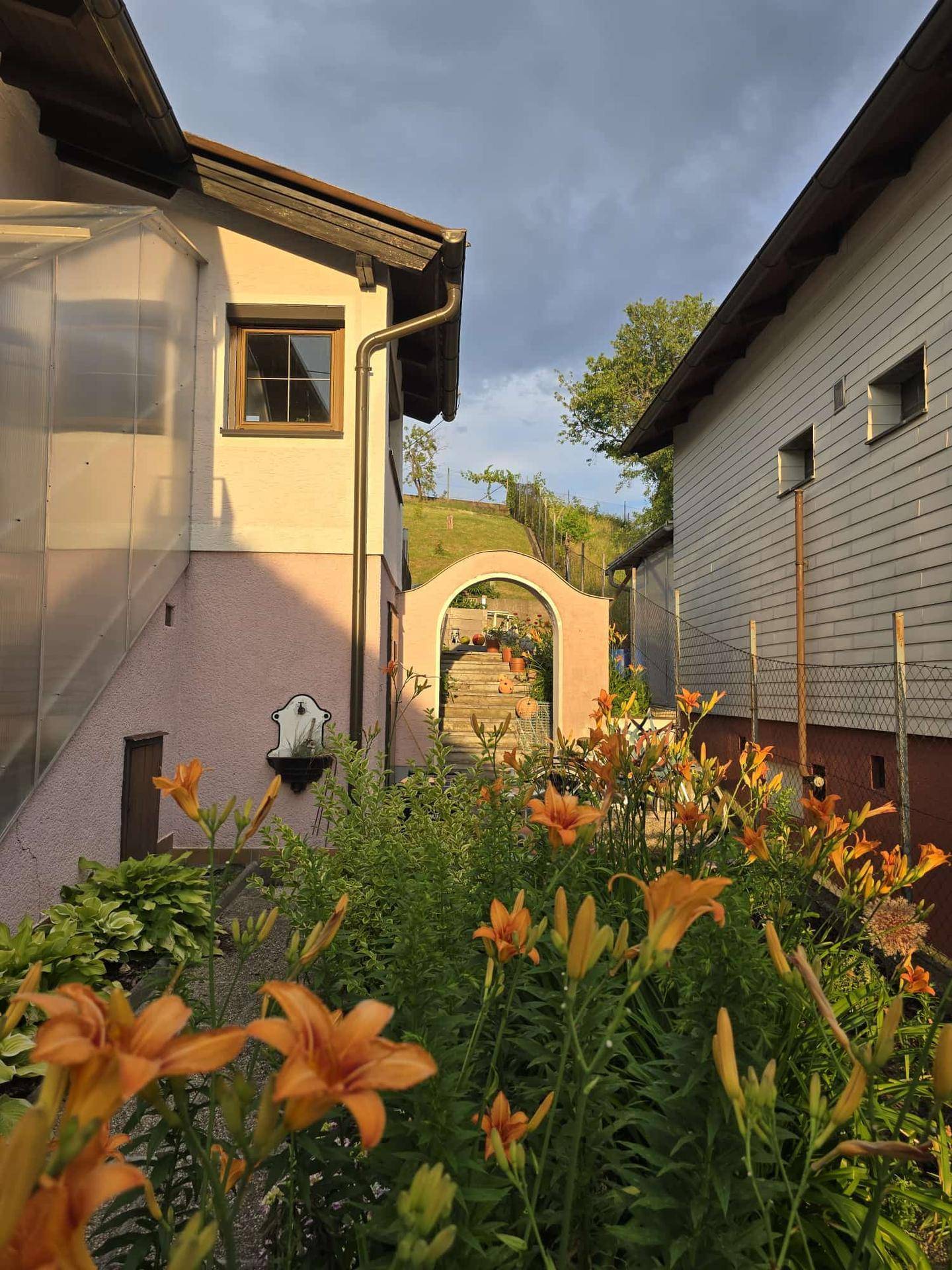 Blick durch einen Gartenweg mit Blumen und einem Torbogen zwischen zwei Häusern bei Sonnenuntergang.