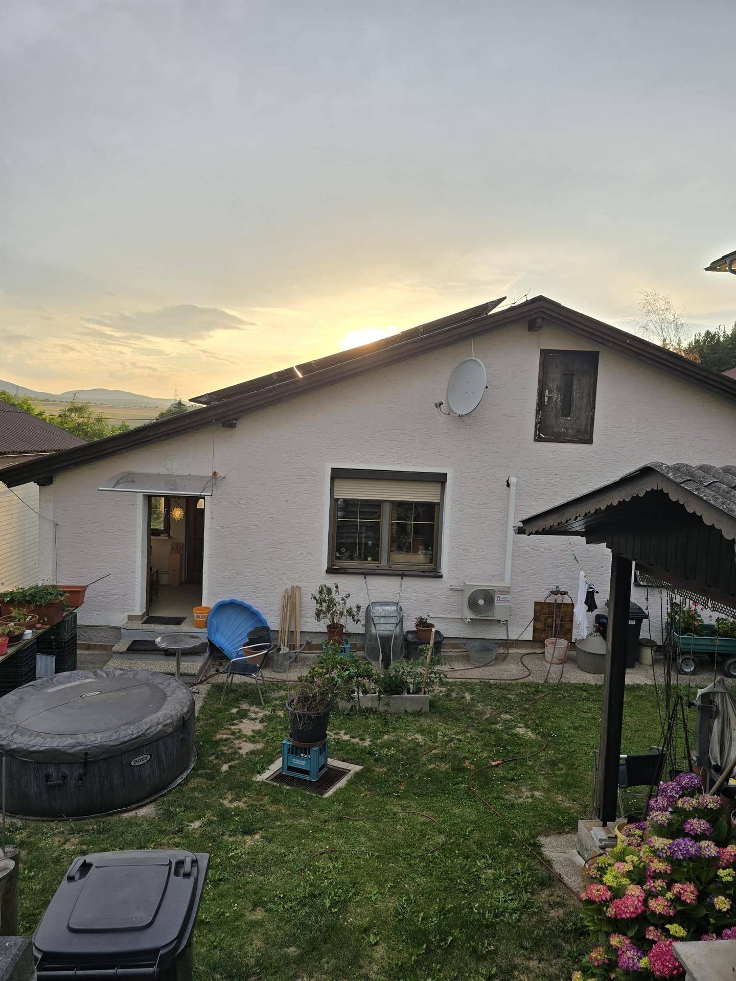 Rückansicht eines weißen Hauses mit Solarpaneelen auf dem Dach und einem Whirlpool im Garten bei Sonnenuntergang.