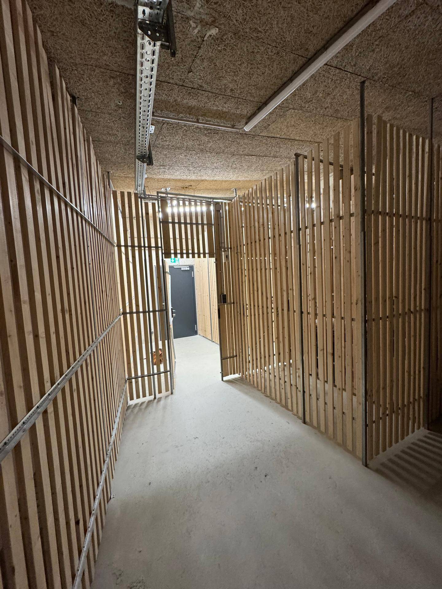 Ein langer Flur in einer Lagereinrichtung mit Holzwänden und einem Betonboden, beleuchtet von Leuchtstoffröhren.