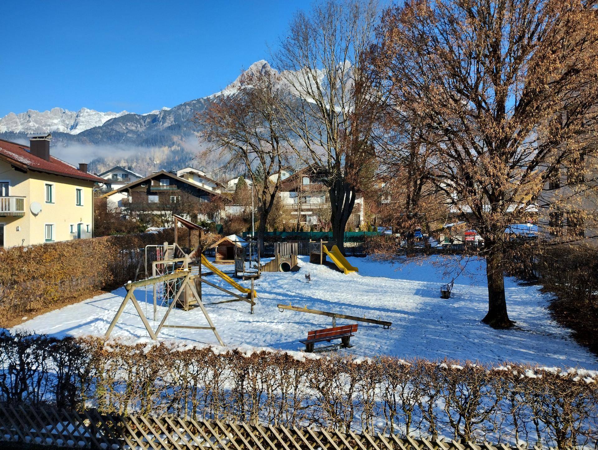 Winterlicher Spielplatz mit Schaukeln und Rutsche, umgeben von Bäumen und mit Bergblick.