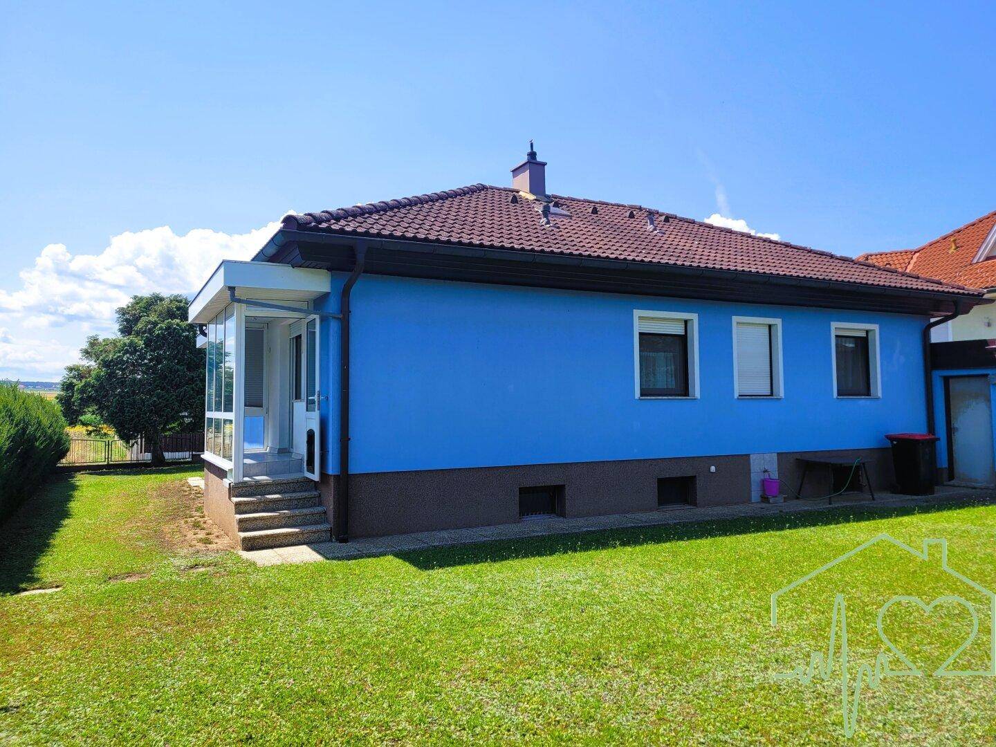 Seitenansicht des Hauses mit blau gestrichener Fassade, Wintergarten und weitläufigem Rasenbereich.