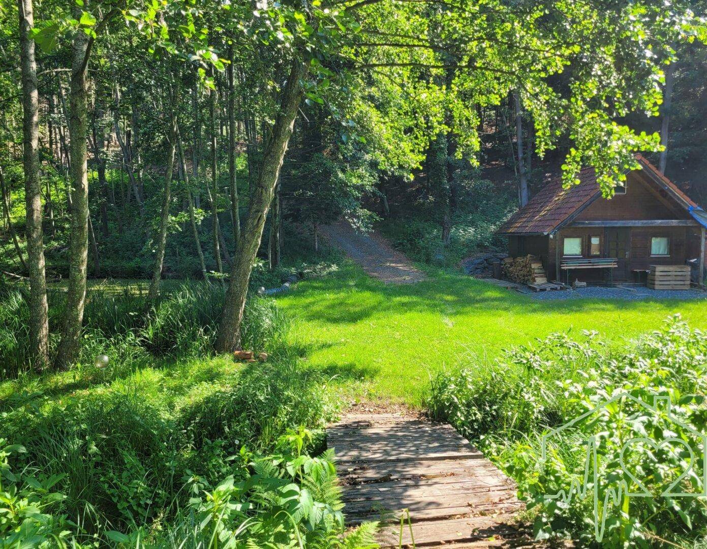 Kleiner Holzsteg über einen Bach, der zu einer idyllischen Holzhütte im Grünen führt.