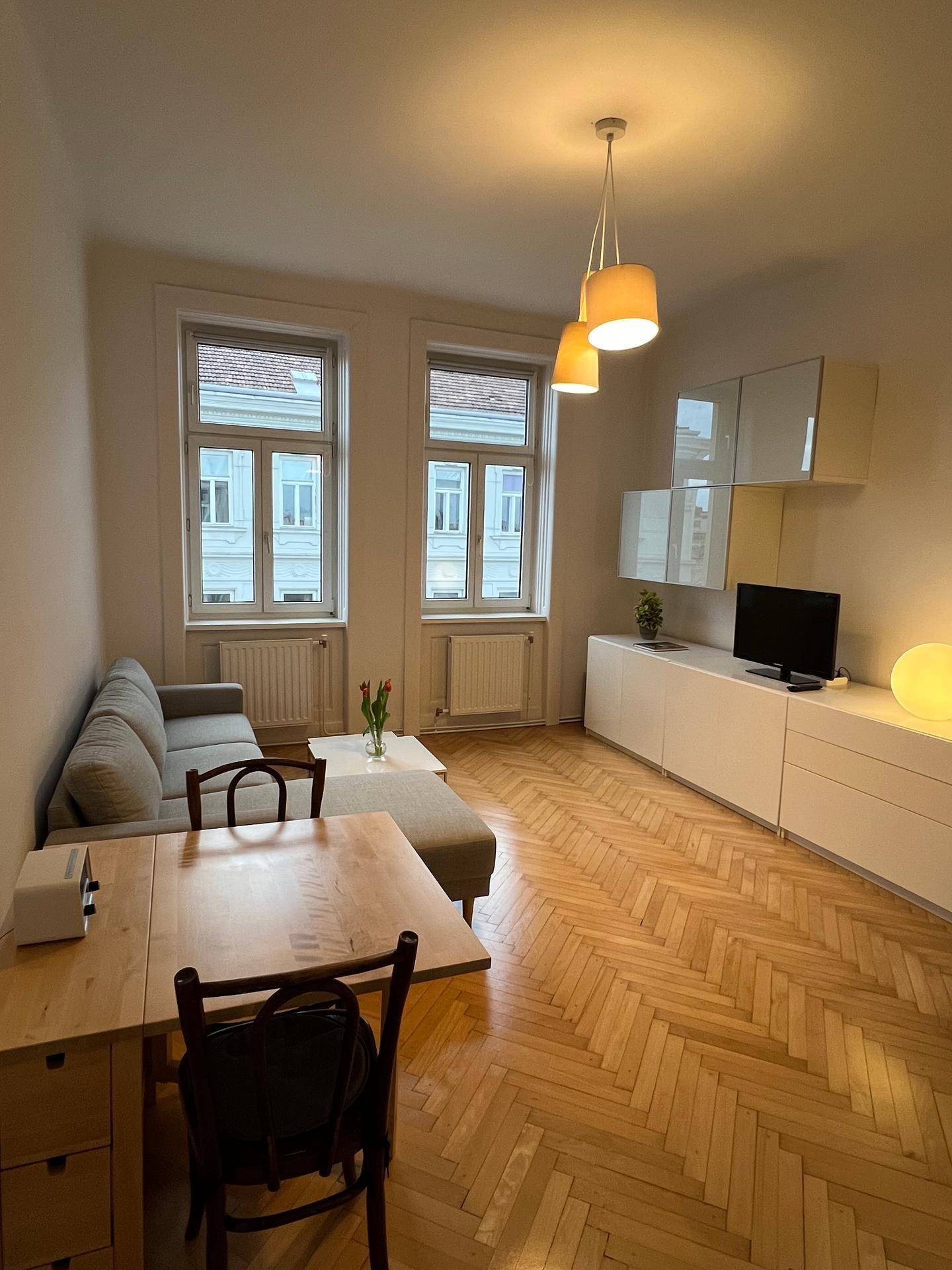 Gemütliches Wohnzimmer mit Esstisch, Sofa und großen Fenstern, die viel Tageslicht hereinlassen.