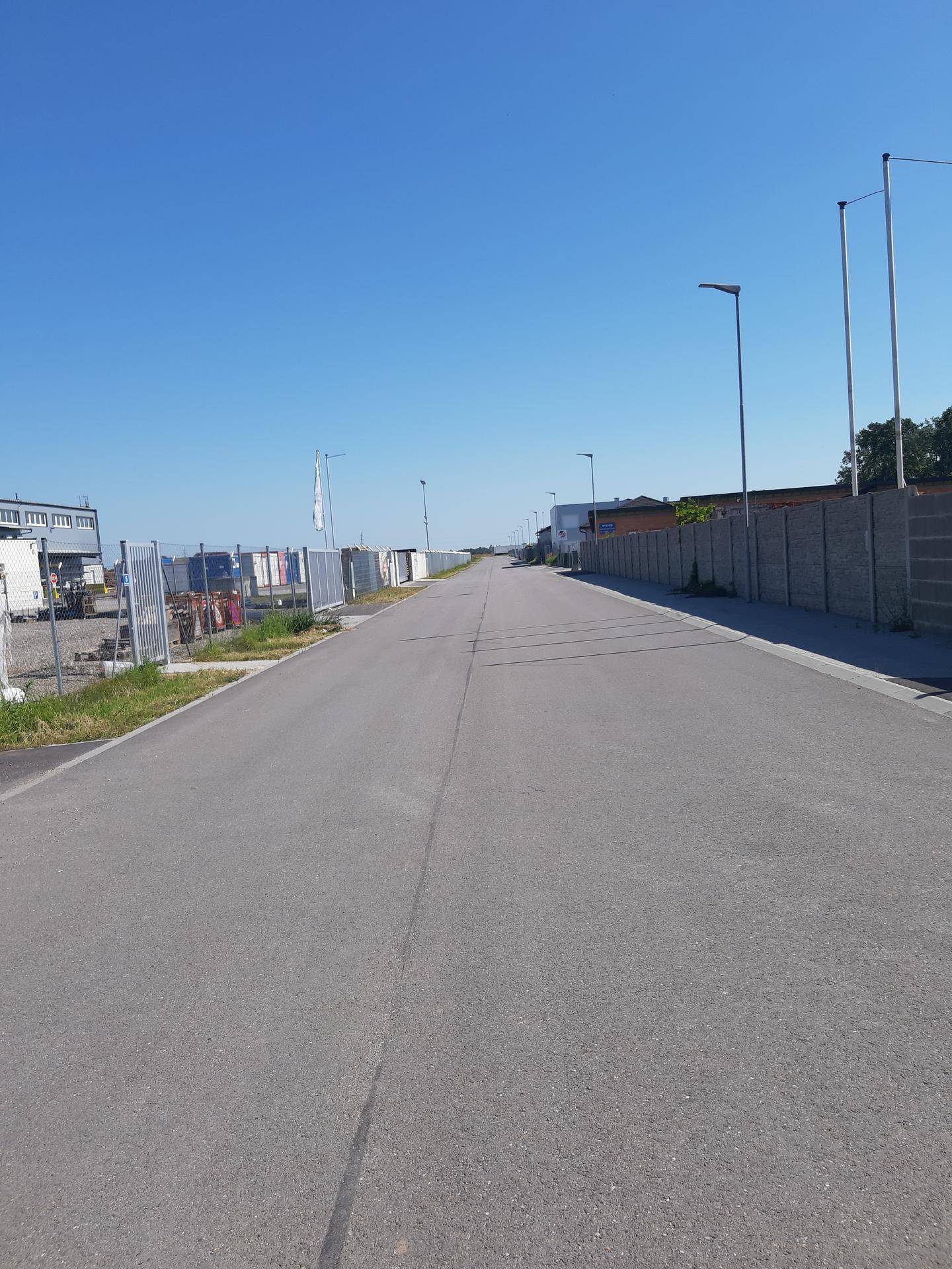 Weitläufige Straße in einem Gewerbegebiet mit Zäunen und Gebäuden unter klarem, blauem Himmel.