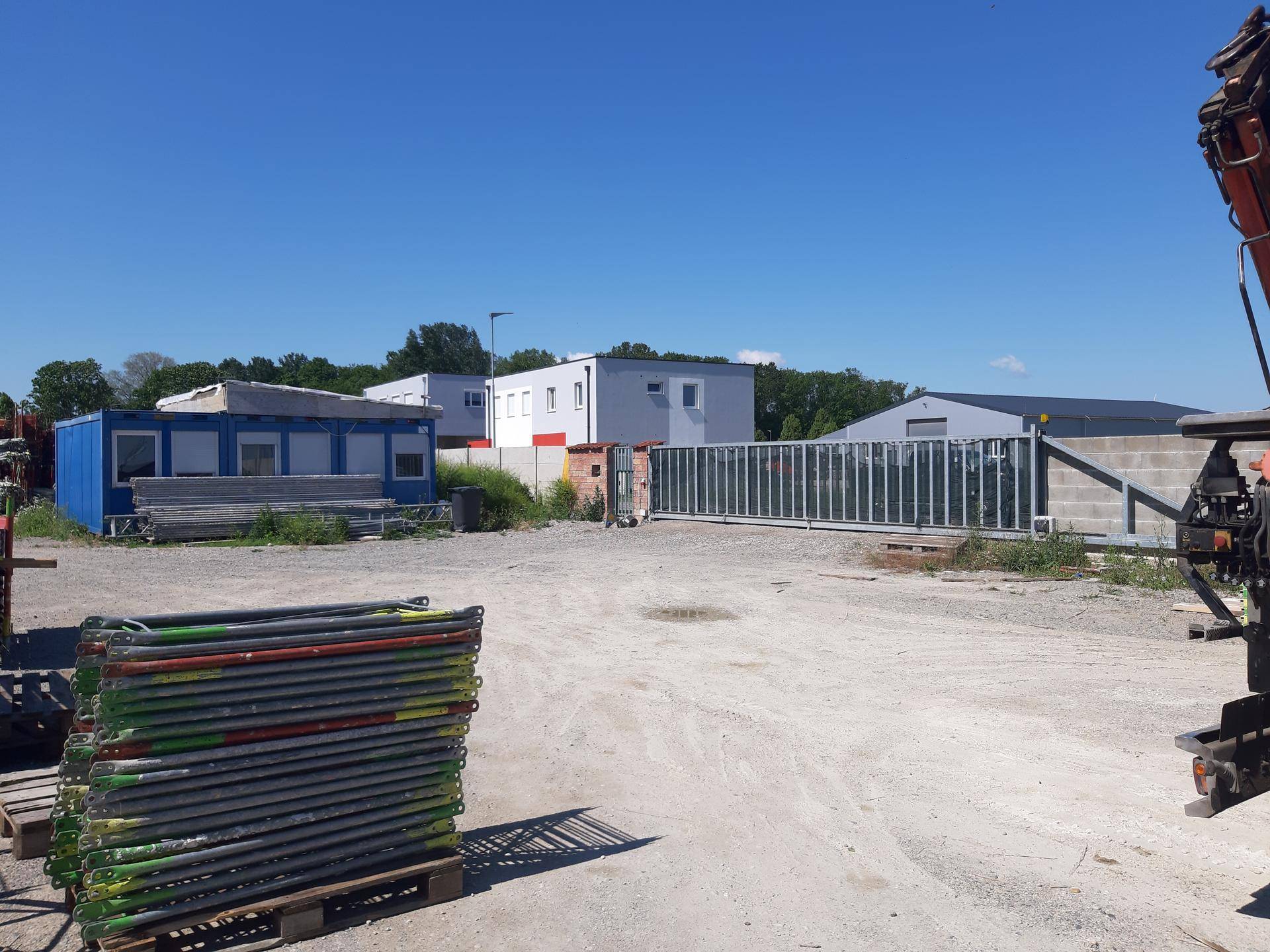 Außenbereich eines Gewerbegrundstücks mit Containern, Gebäuden und einem Schotterplatz unter blauem Himmel.