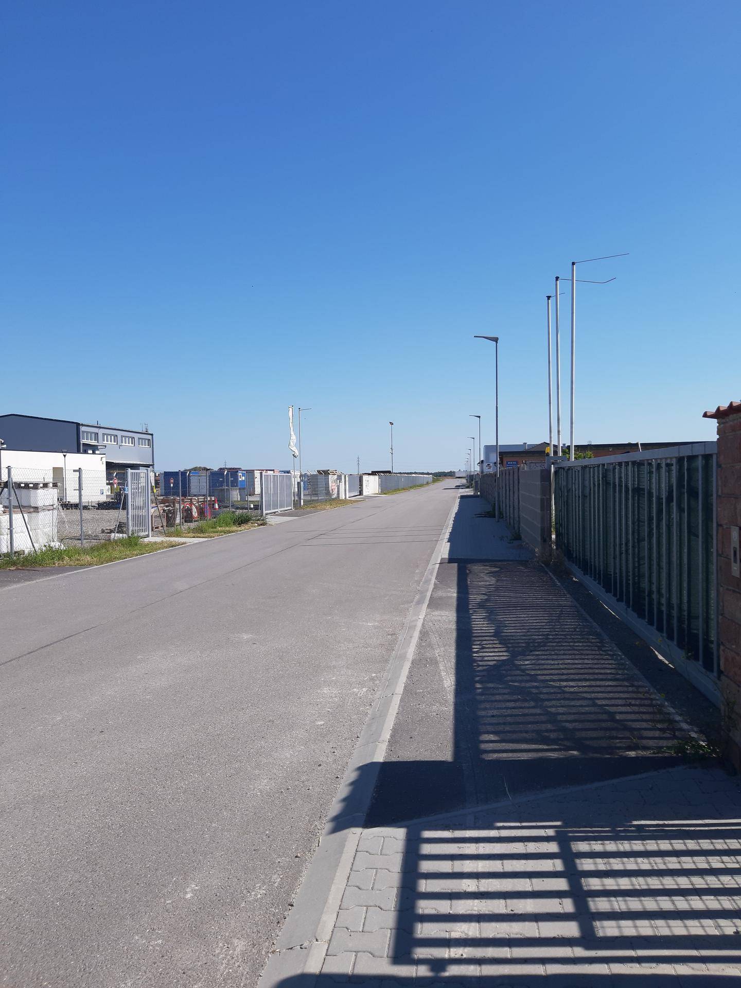 Straße mit Bürgersteig und Metallzäunen, die zu einem Industriegebiet führen, unter klarem blauem Himmel.