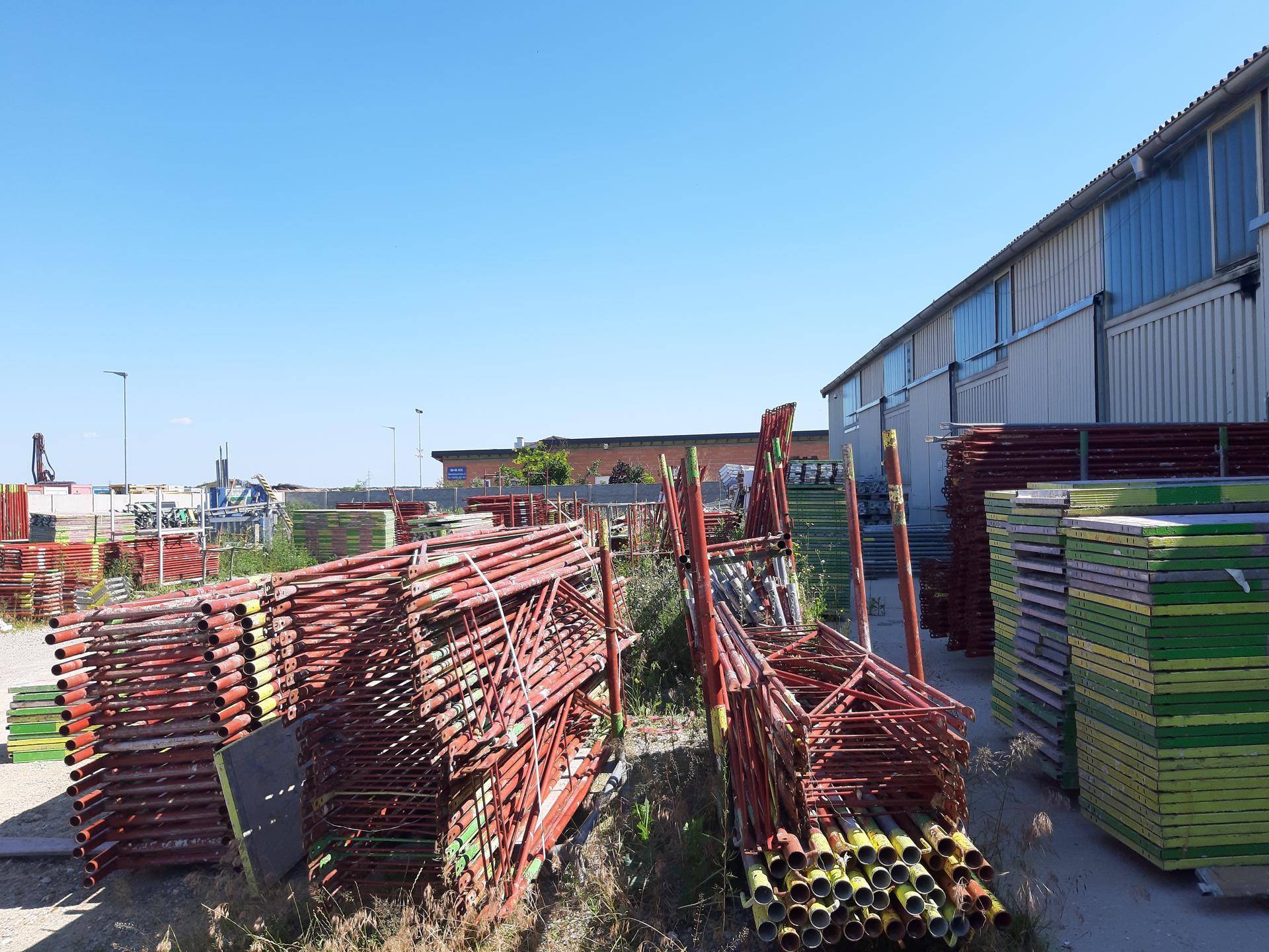 Außenlagerbereich mit gestapelten Gerüstteilen und einem Industriegebäude im Hintergrund unter blauem Himmel.