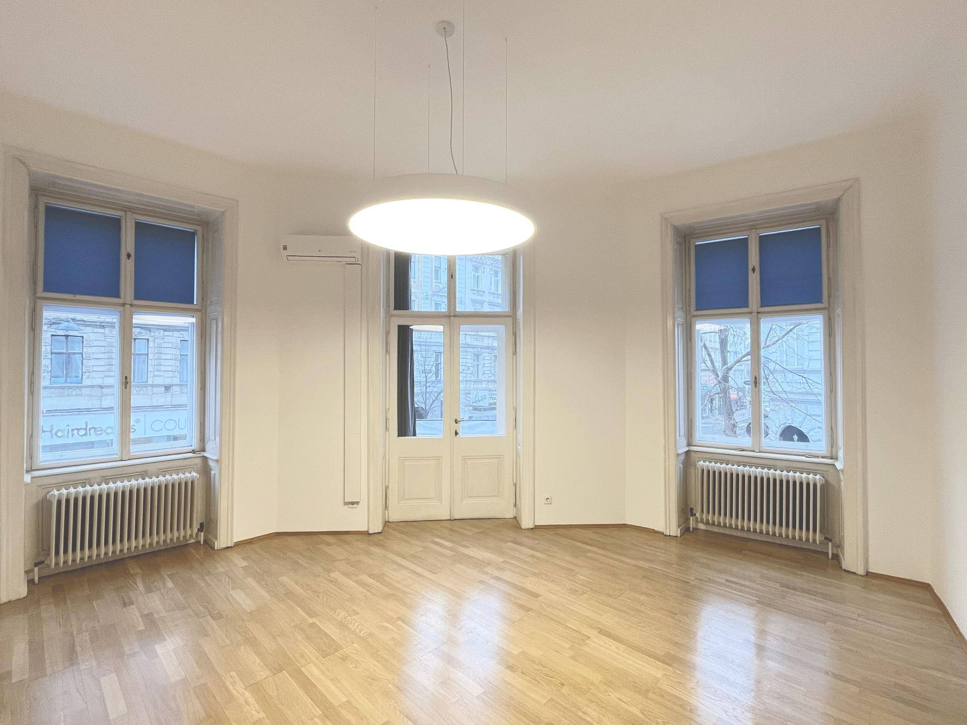 Geräumiger Wohnbereich mit Parkettboden, Klimaanlage und hohen Fenstern, die viel Tageslicht hereinlassen.