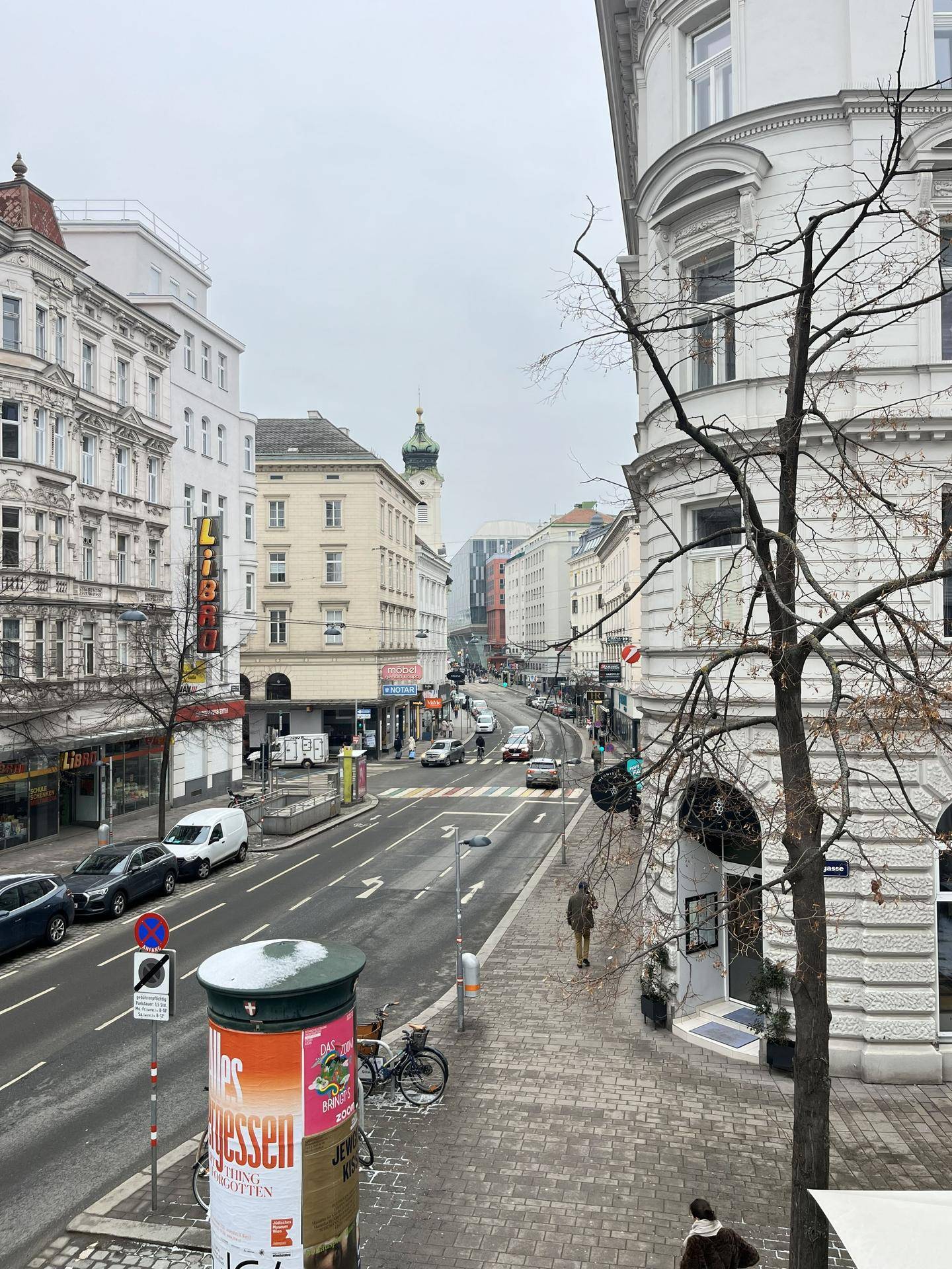 Blick aus dem Fenster auf eine belebte Stadtstraße mit Gebäuden und Fahrzeugen.
