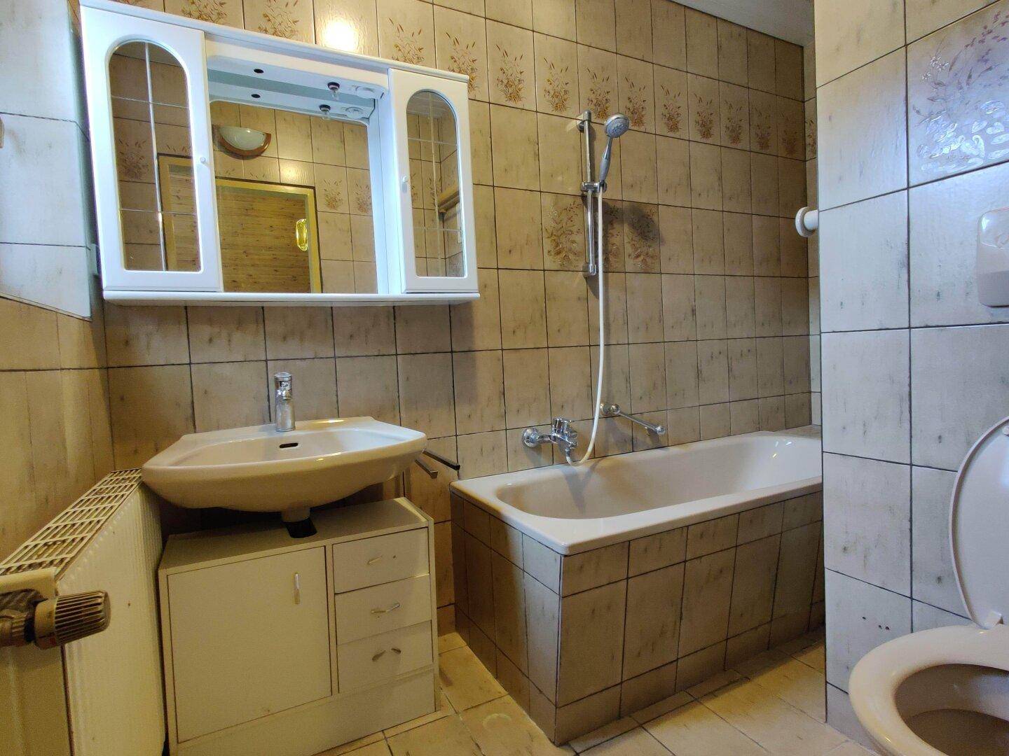 Badezimmer mit Badewanne, Waschbecken und Spiegelschrank, gefliest in beigen Tönen.