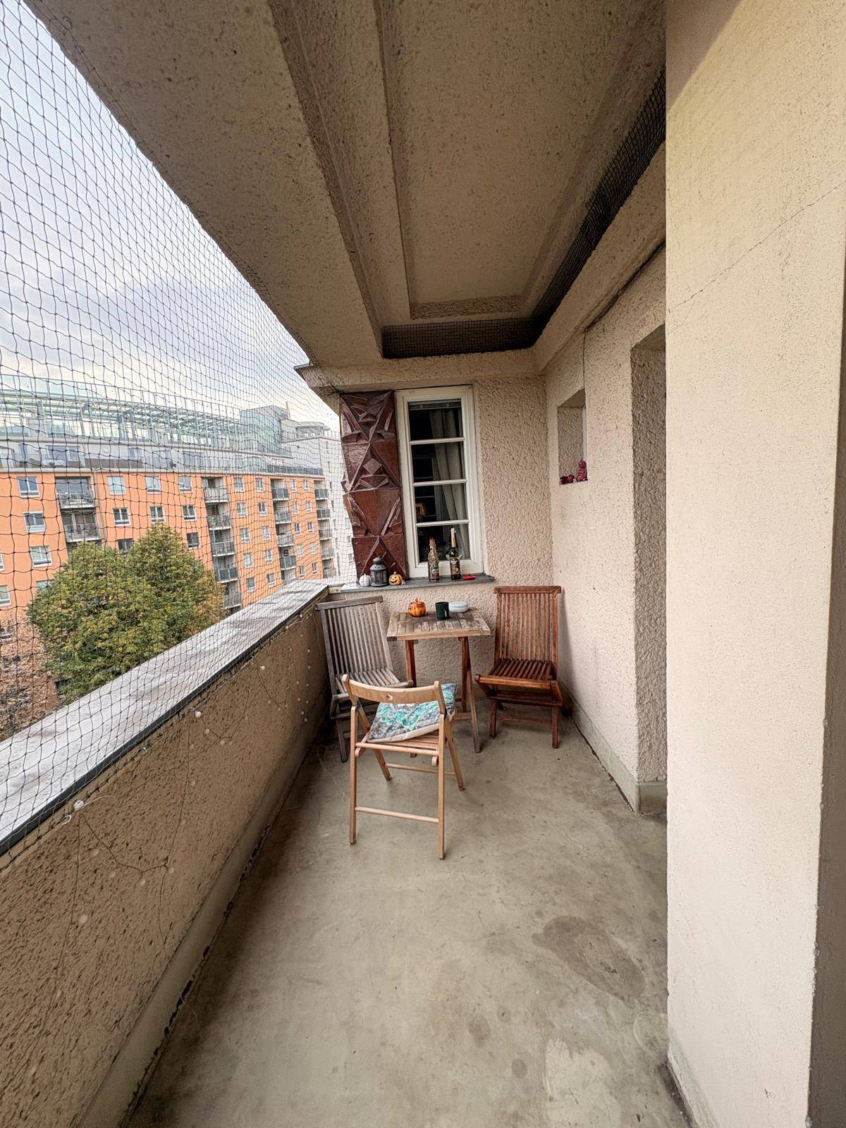 Balkon mit Holztisch und Stühlen, umgeben von einem Sicherheitsnetz, bietet Blick auf die Stadt.