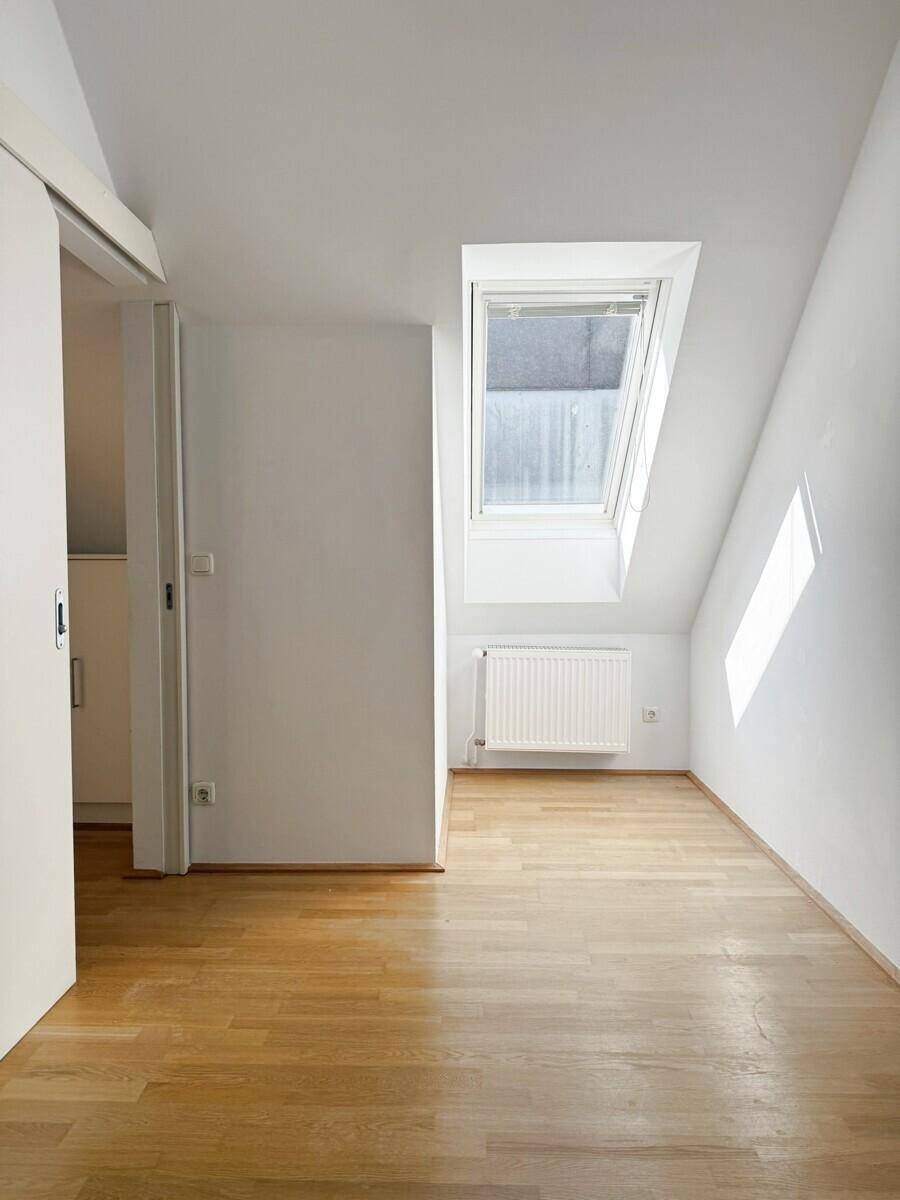Dachzimmer mit schräger Decke, hellem Parkettboden, Dachfenster und Heizkörper.