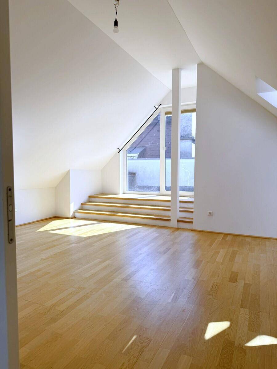 Helles Dachzimmer mit schrägen Wänden, Holzboden und einem großen Fensterbereich mit Stufen.
